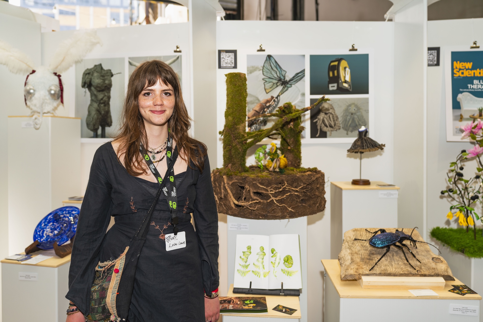 A woman with long brown hair stands in front of an exhibition. She wears a black dress and a lanyard. Behind her is an array of art pieces, including sculptures of insects and a blue glass object on a nearby display. The setting is well-lit with white walls.