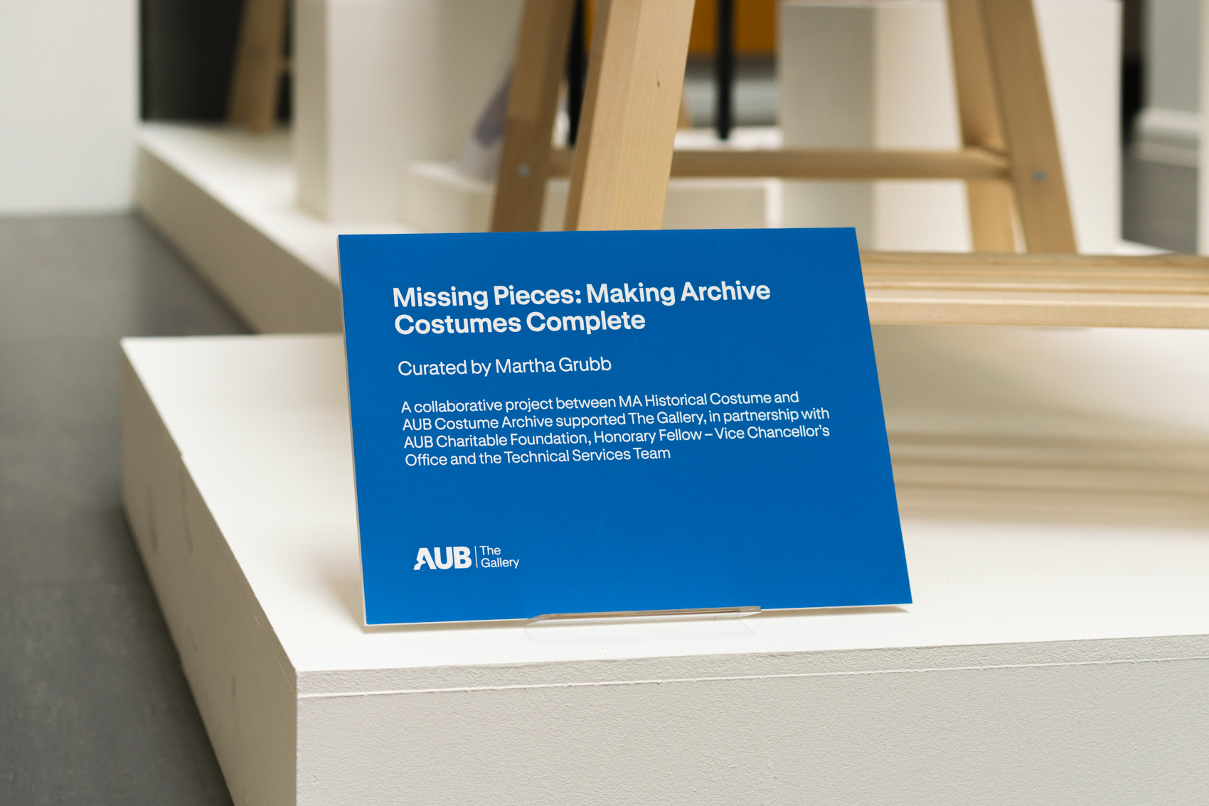 Blue plaque with text that reads 'Missing Pieces: Making Archive Costumes Complete, Curated by Martha Grubb, A collaborative project between MA Historical Costume and AUB Costume Archive supported The Gallery, in partnership with AUB Charitable Foundation, Honorary Fellow - Vice Chancellor's Office and the Technical Services Team'.