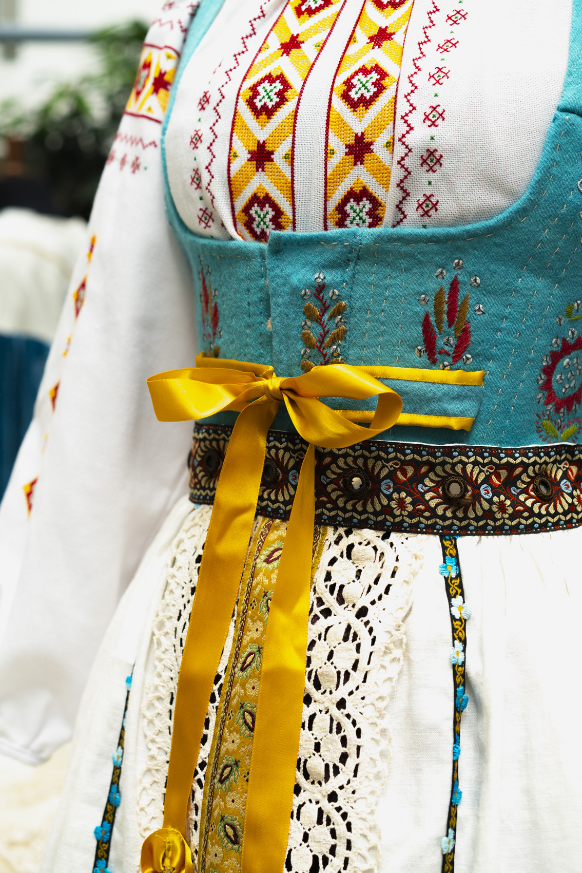 Close-up of detailed dress with red-yellow pattering, teal corset piece and golden ribbon.