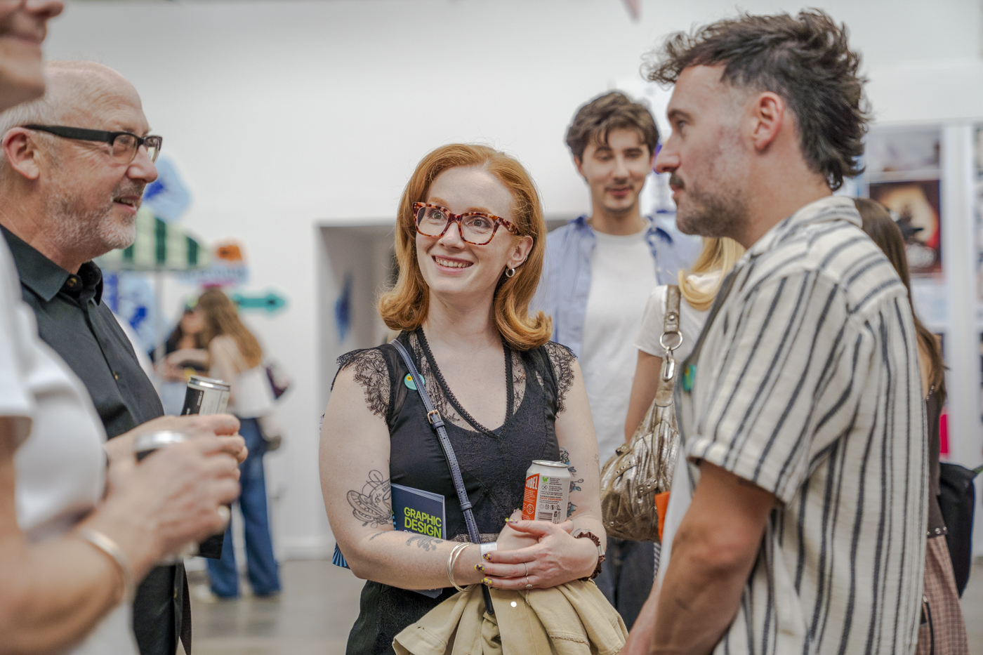 A red-haired woman with glasses stands with a drink cup and book, talking with three people in a bright indoor space.