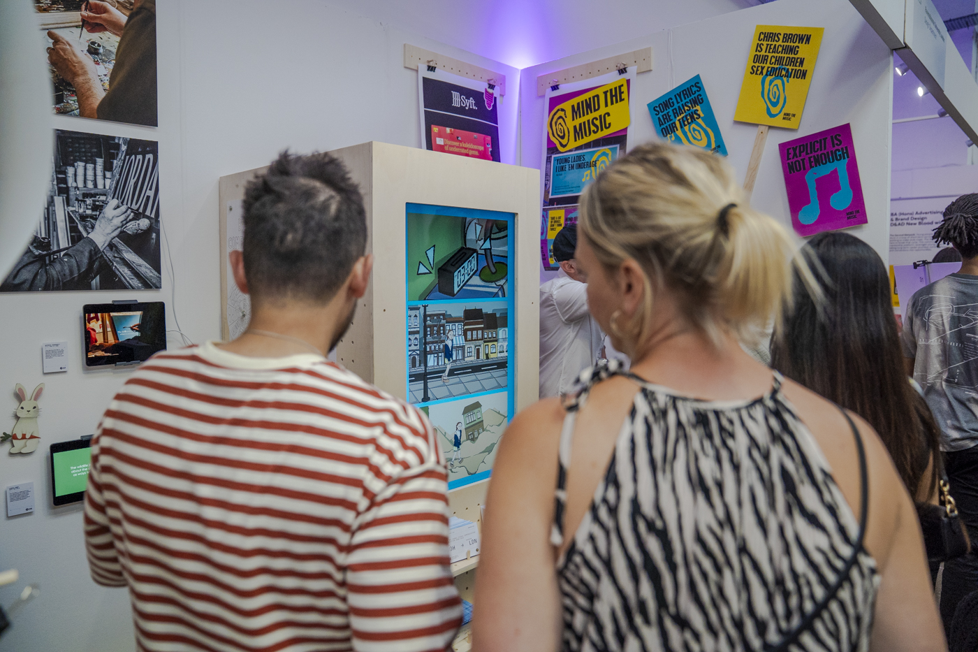 Three people view digital art displays and colourful posters in a gallery space with white walls and purple lighting.