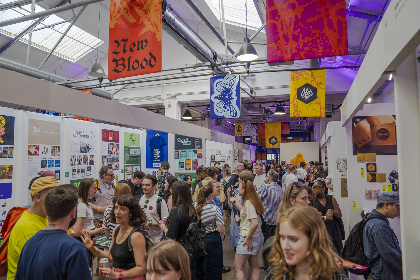 A crowded art fair with visitors standing between white display walls and hanging banners beneath a bright industrial ceiling.