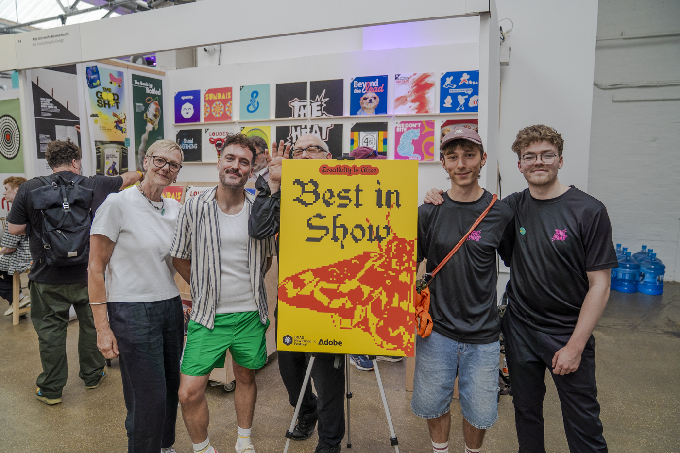 A group of five people stand indoors holding a yellow and red “Best in Show” poster on an easel, with other colourful art prints displayed on the wall behind them.