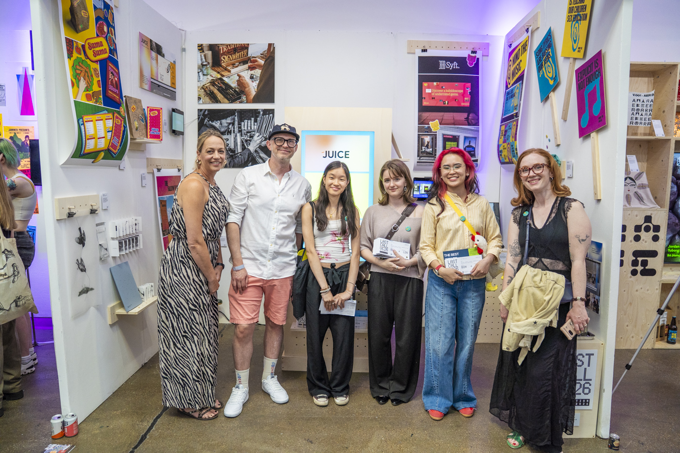 Six people stand together at an exhibition booth, with colourful artwork, posters and a lit display reading "JUICE" in the background.
