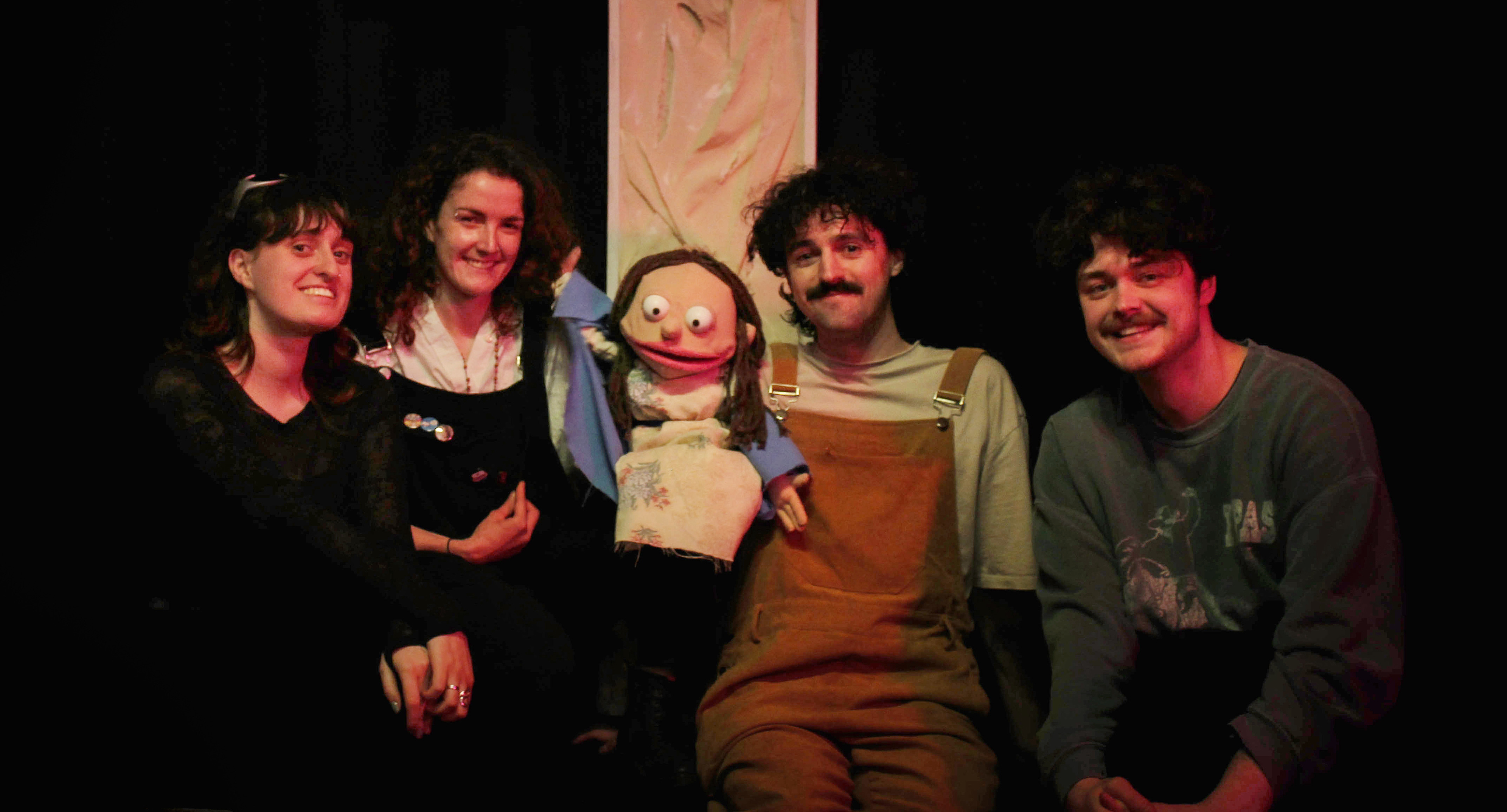 A group of directors and actors from Kayleigh Benham's play 'Saint', posed with a puppet in front of a cross in red lighting, black background.