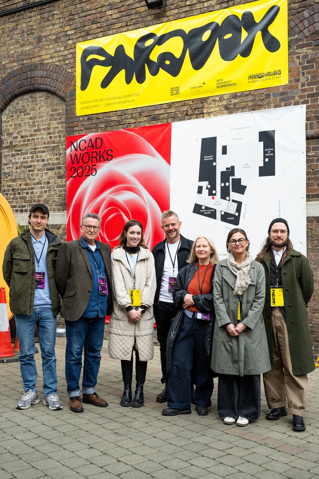 Seven people standing in front of brick building with yellow banner reading 'SHREDDER' and white banner with red spiral design reading 'NGAD WORKS 2030'