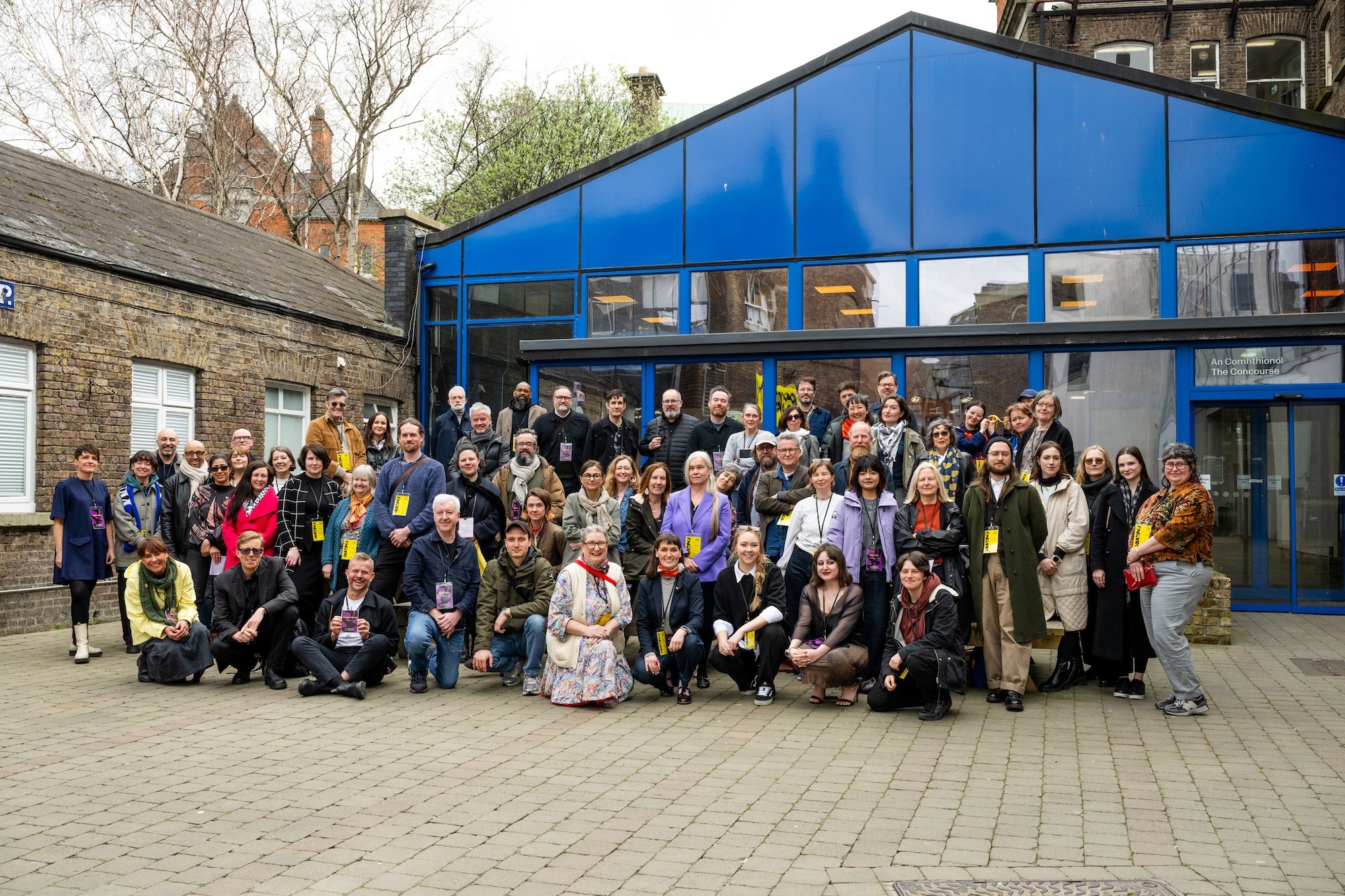 Large group of approximately 60 people posing together outside blue modern building with glass front and traditional stone buildings behind