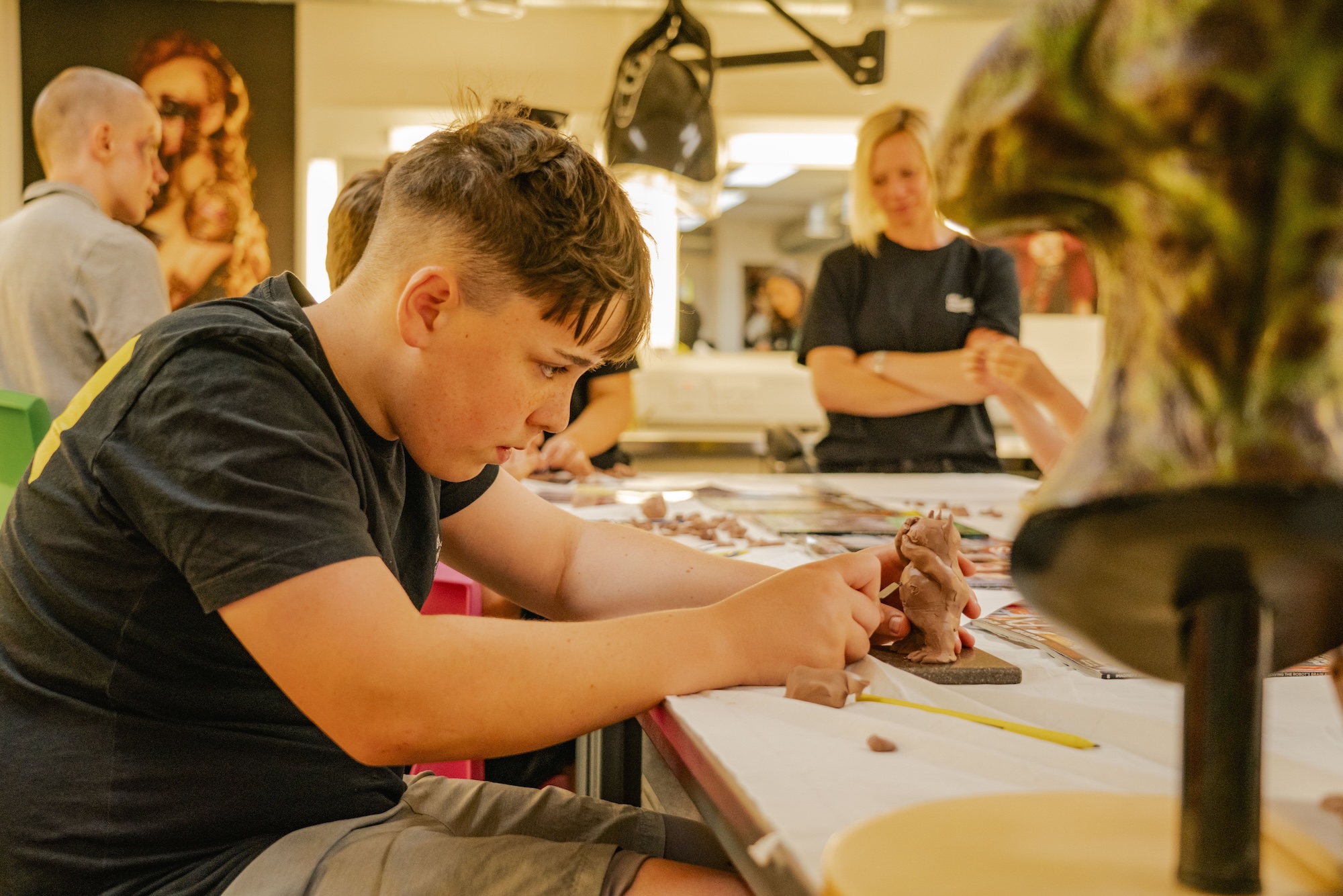 A young person focused on sculpting a clay figure at a work table in a workshop. In the background, two adults stand, and a sculpture is partially visible on the right side of the image.