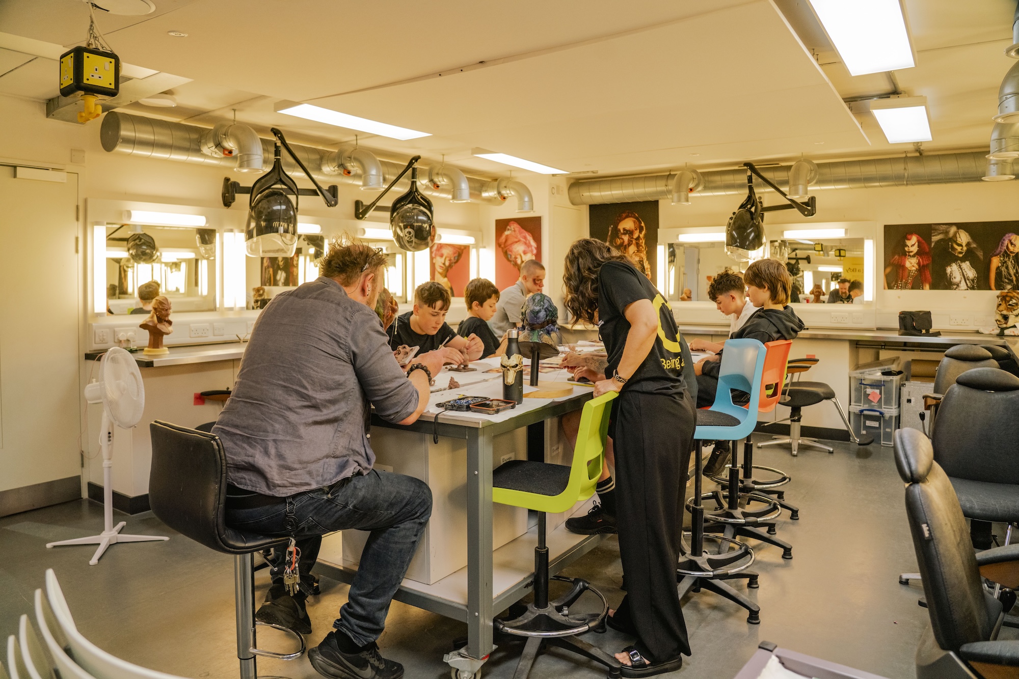 A bright makeup studio with several people gathered around a central table. Two individuals are seated, while others are engaged in activities at the table. Mirrors are visible along the walls, and makeup supplies are scattered across the workspace.