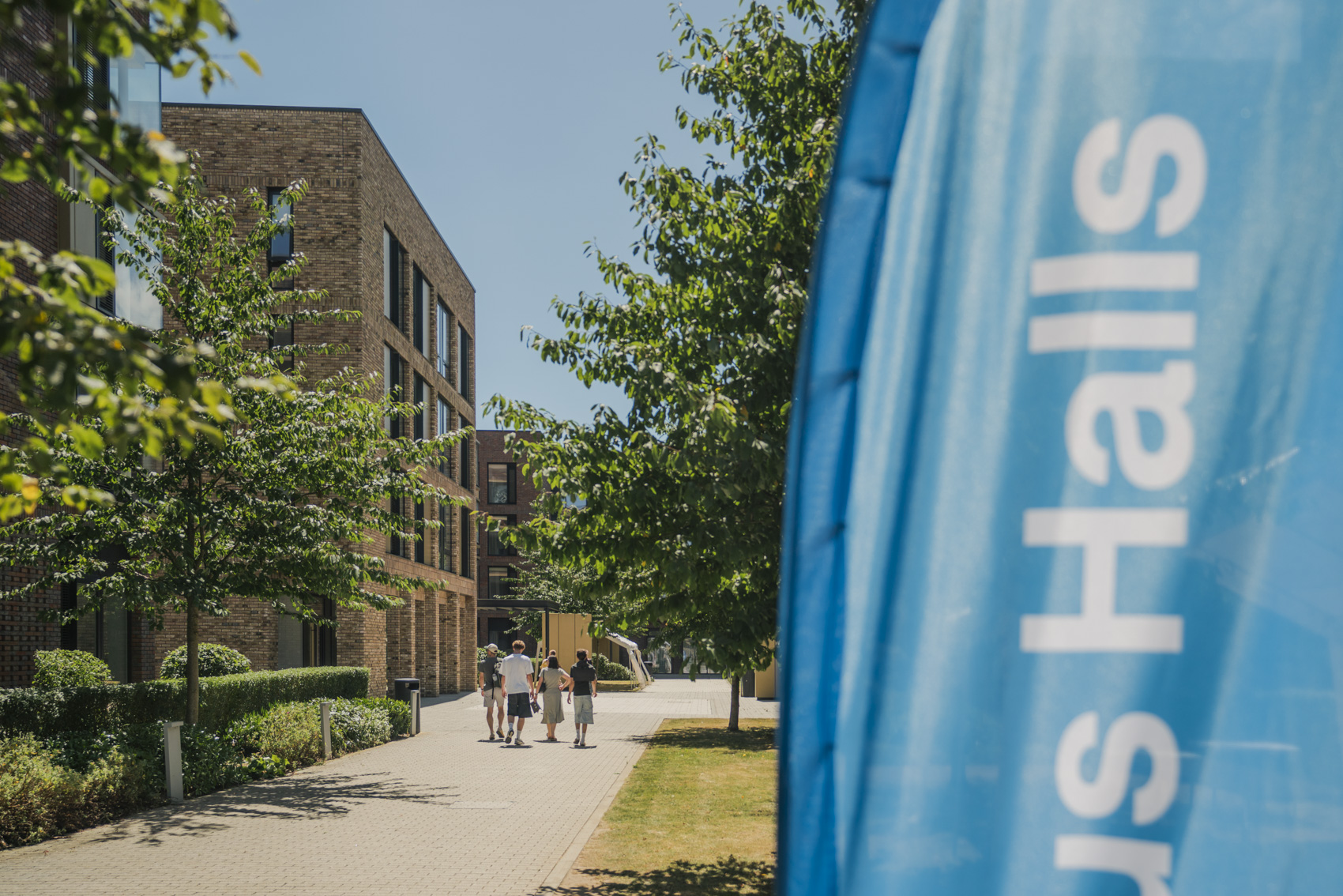 A blue vertical banner reading Hills Halls stands beside a tree-lined campus walkway with several people walking past brick buildings.
