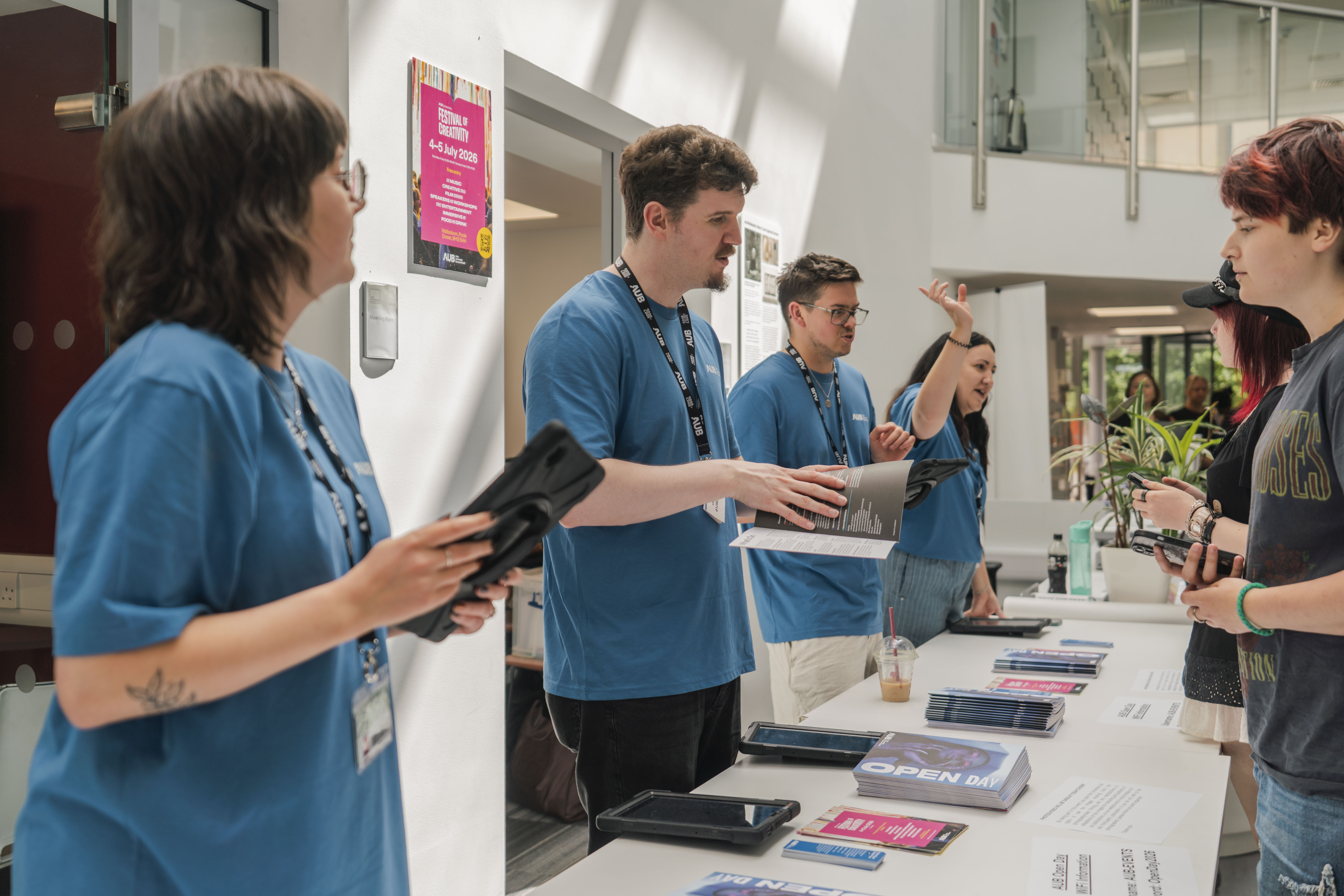 A group of staff in blue T-shirts stands behind a table of brochures and tablets, speaking with two visitors in a bright lobby.