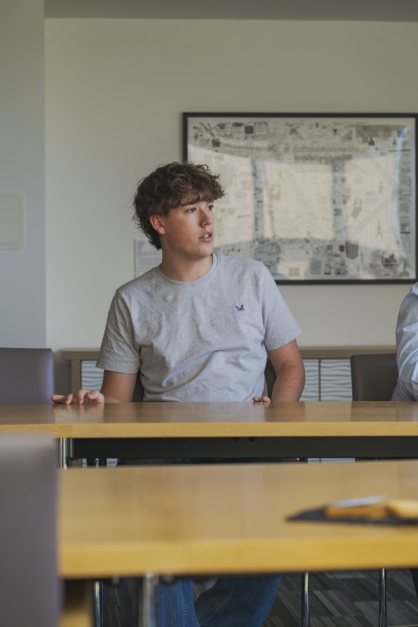 A young man with curly hair wearing a grey T-shirt sits at a long wooden table in a modern room. A framed map is visible on the wall behind him, and another person is seated beside him, partially out of view.