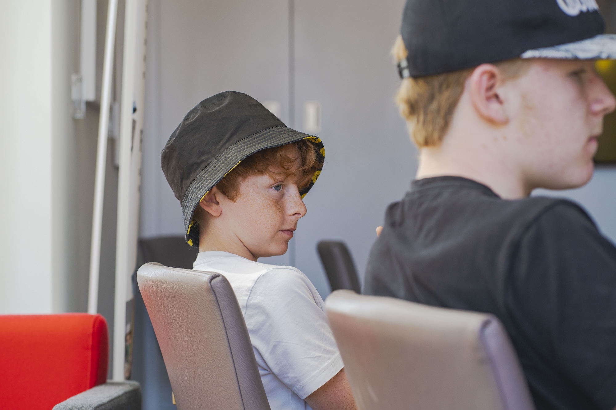 A boy with curly hair wearing a black bucket hat and a white T-shirt is seated on the left, looking to the side. Another young man with a baseball cap and light brown hair is visible in profile on the right, both seated in contemporary chairs.