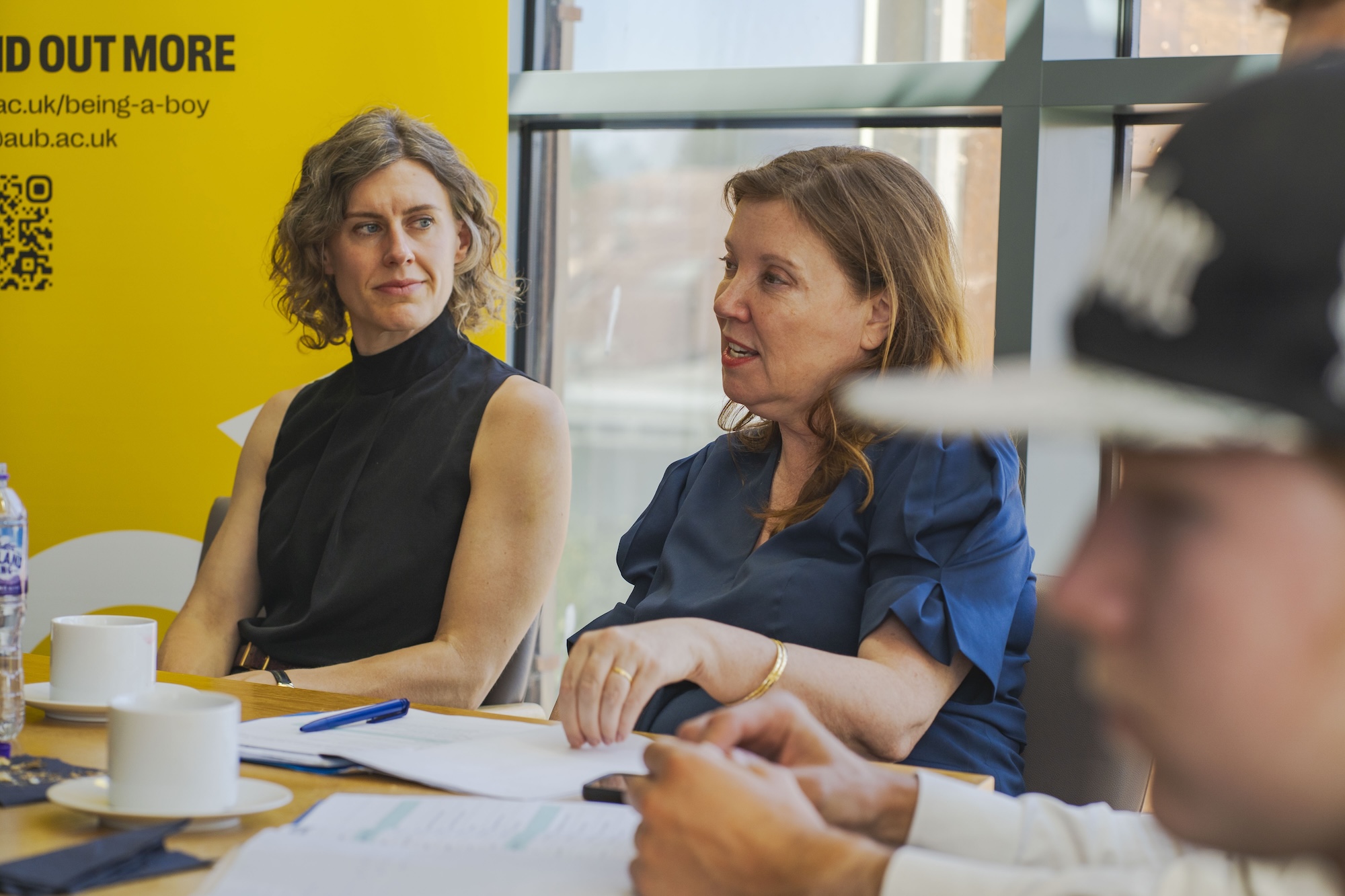 J Toale MP and Dame R de Souza sit at a table in a meeting room. Papers and stationary are visible on the table.