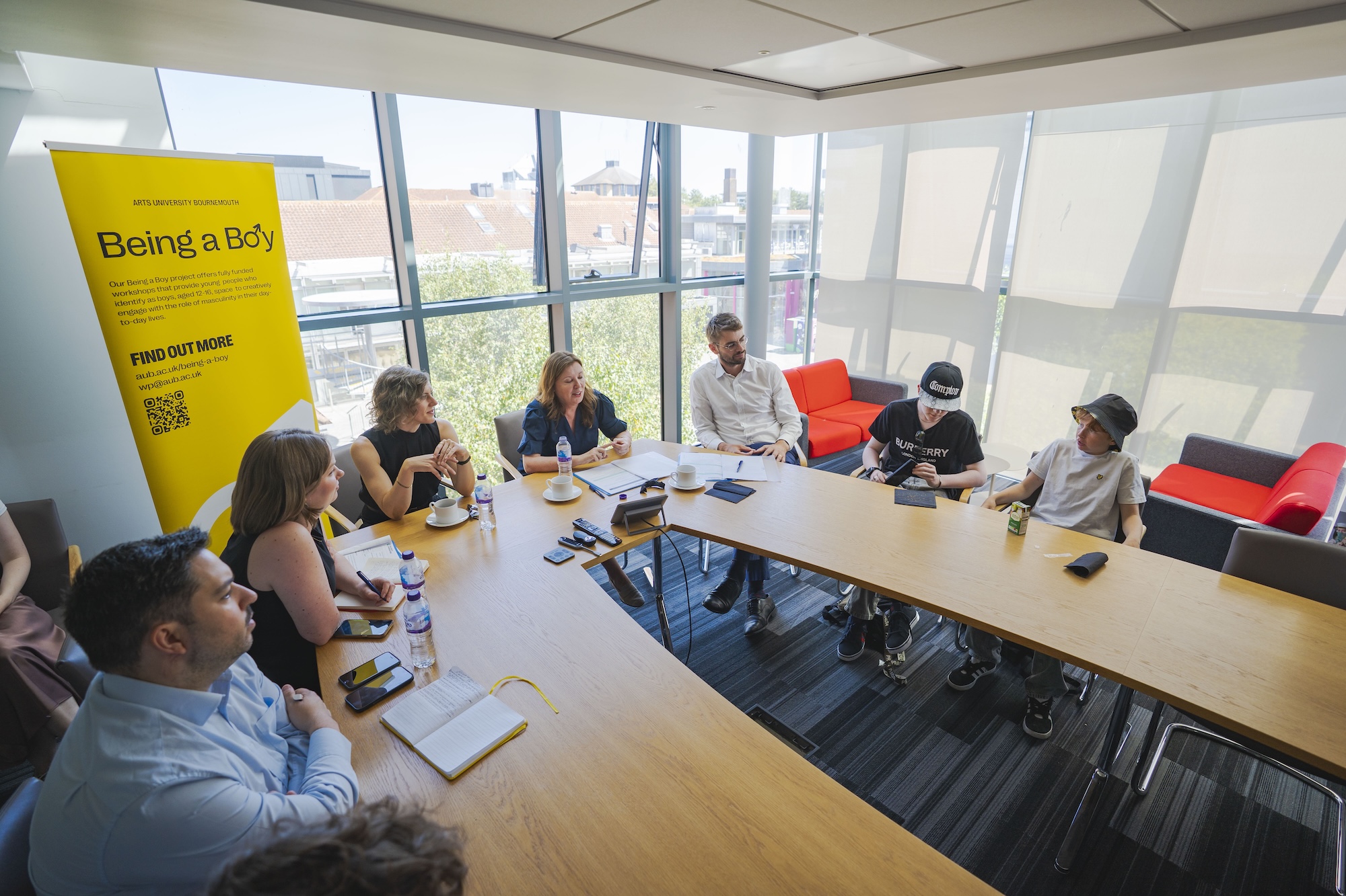 A group of eight people are seated around a long wooden table in a brightly lit meeting room with large windows. One person is wearing a black hat and another has a white blindfold. A yellow poster titled 'Being a Body' is displayed on the wall.