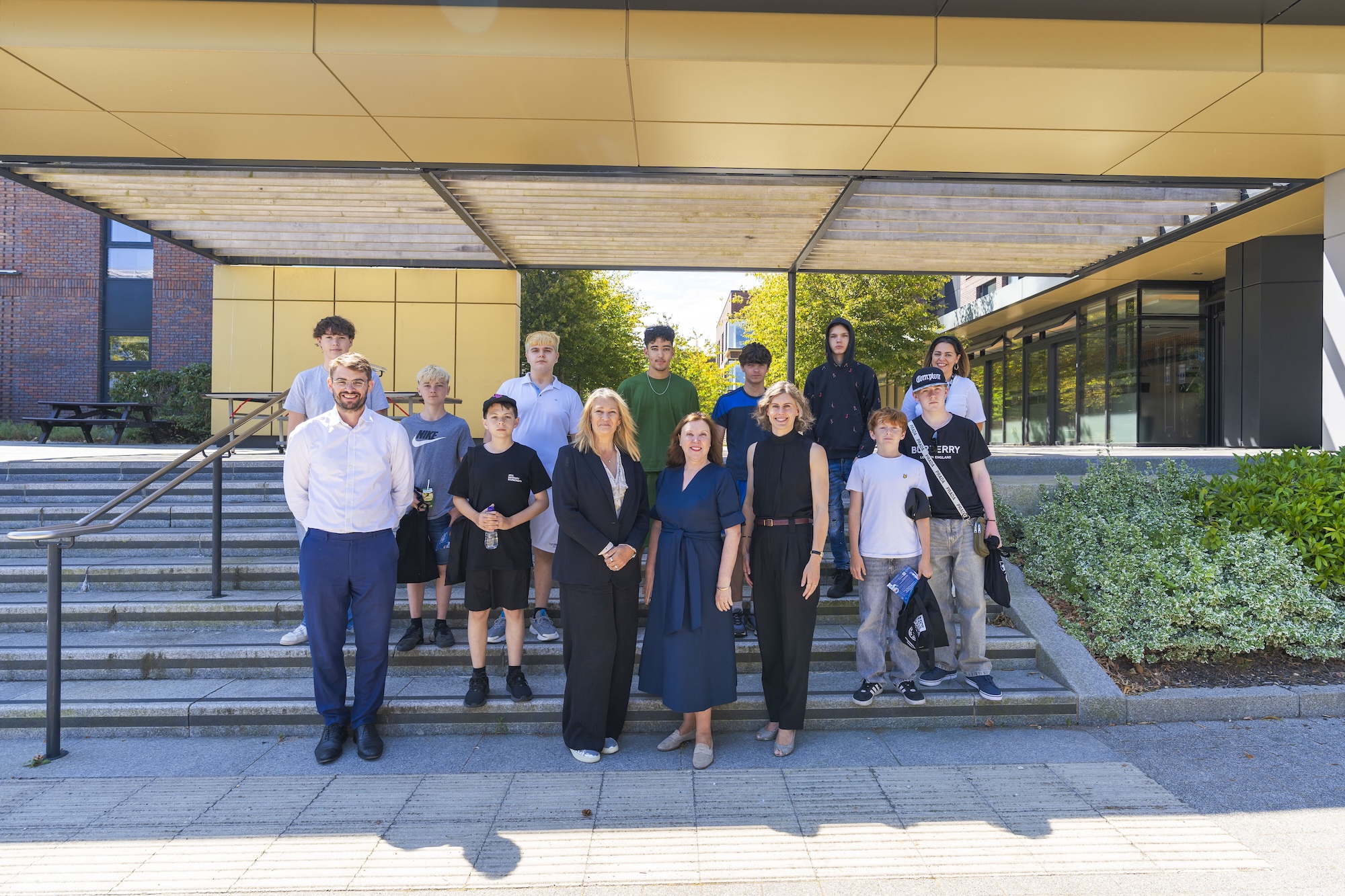 A group of 11 individuals, including students and adults, stands on steps in front of a modern building with large windows and greenery. The adults are dressed in formal attire, while the students wear casual clothing.