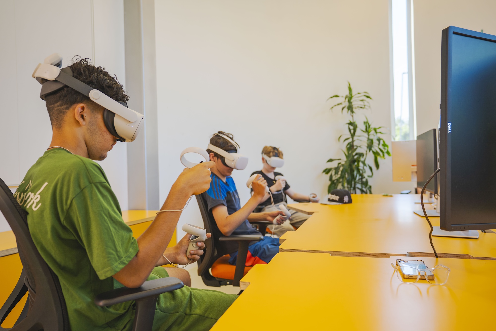 Three young individuals are seated at a long orange table, wearing virtual reality headsets and holding controllers. The room features large windows with greenery visible outside, and there is a potted plant in the background.