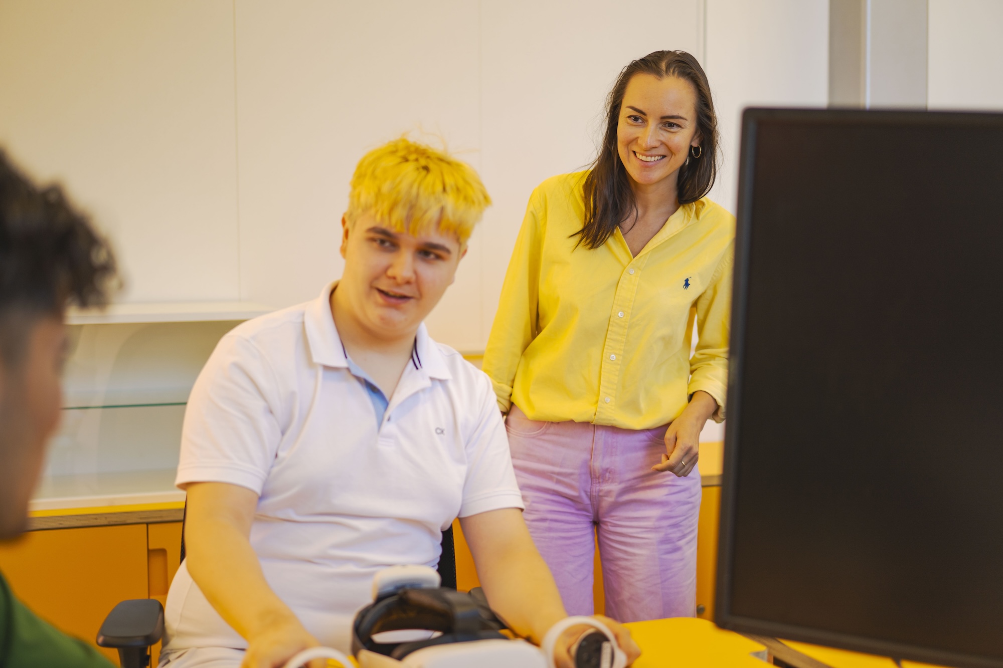 A young man with short blonde hair, wearing a white polo shirt, sits at a desk with a virtual reality headset. A woman in a yellow shirt stands next to him, observing. A computer monitor and orange cabinetry are visible in the background.
