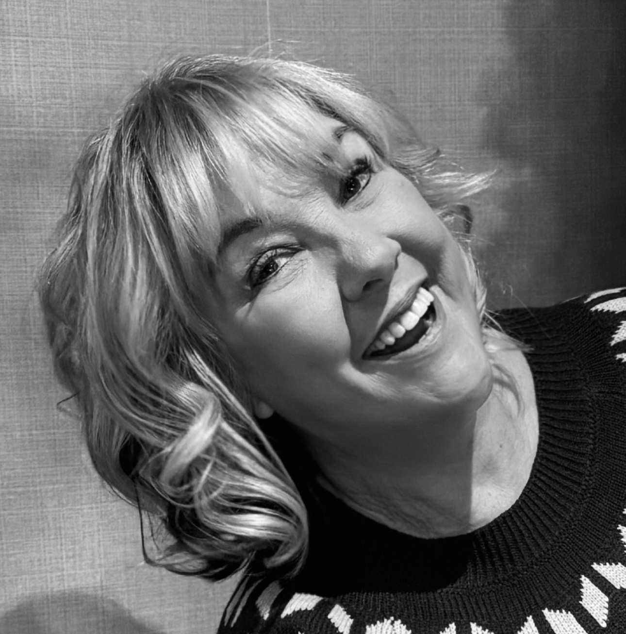 Black and white portrait of woman with blonde bob haircut and fringe, smiling broadly whilst tilting her head, wearing dark clothing.