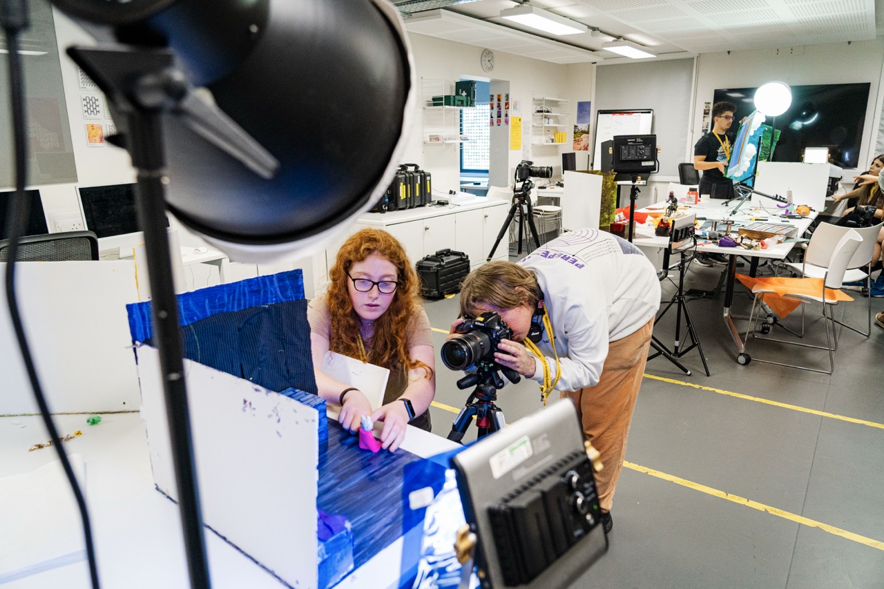 Two people work with a camera and lighting equipment in a studio, with tables, monitors and cables around them.