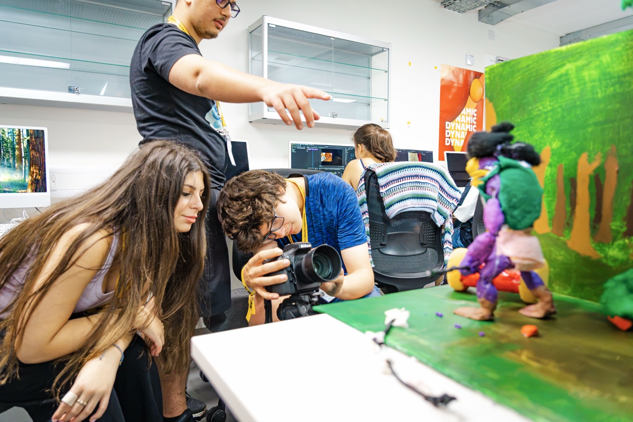 A group of students work around a camera filming a small colourful stop-motion scene in a classroom or studio, with a green backdrop and props on a table.