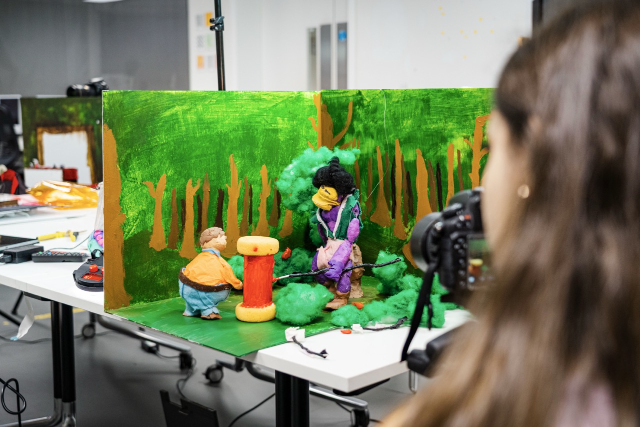 A photographer films a small handmade forest diorama with two figurines on a table in a studio.