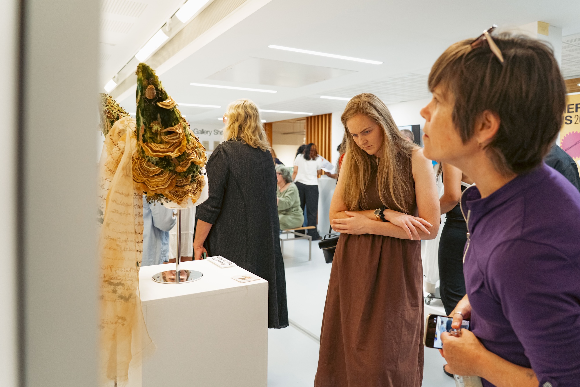A woman in a purple top stands beside a blonde woman in a brown dress, with a displayed textile sculpture on a pedestal in a bright gallery space.