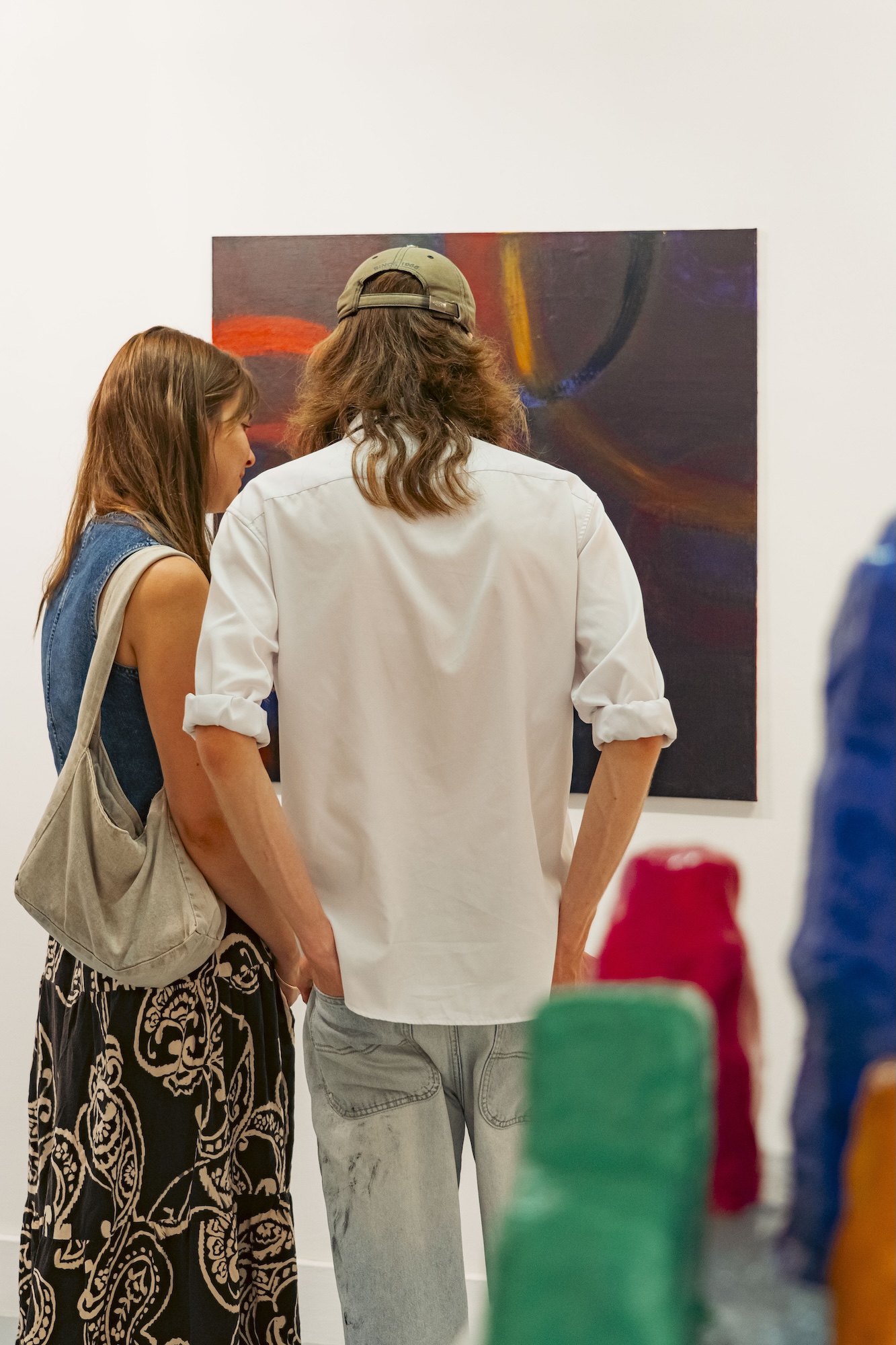 A person in a white shirt and cap and a person with a shoulder bag stand in front of an abstract painting in a gallery.