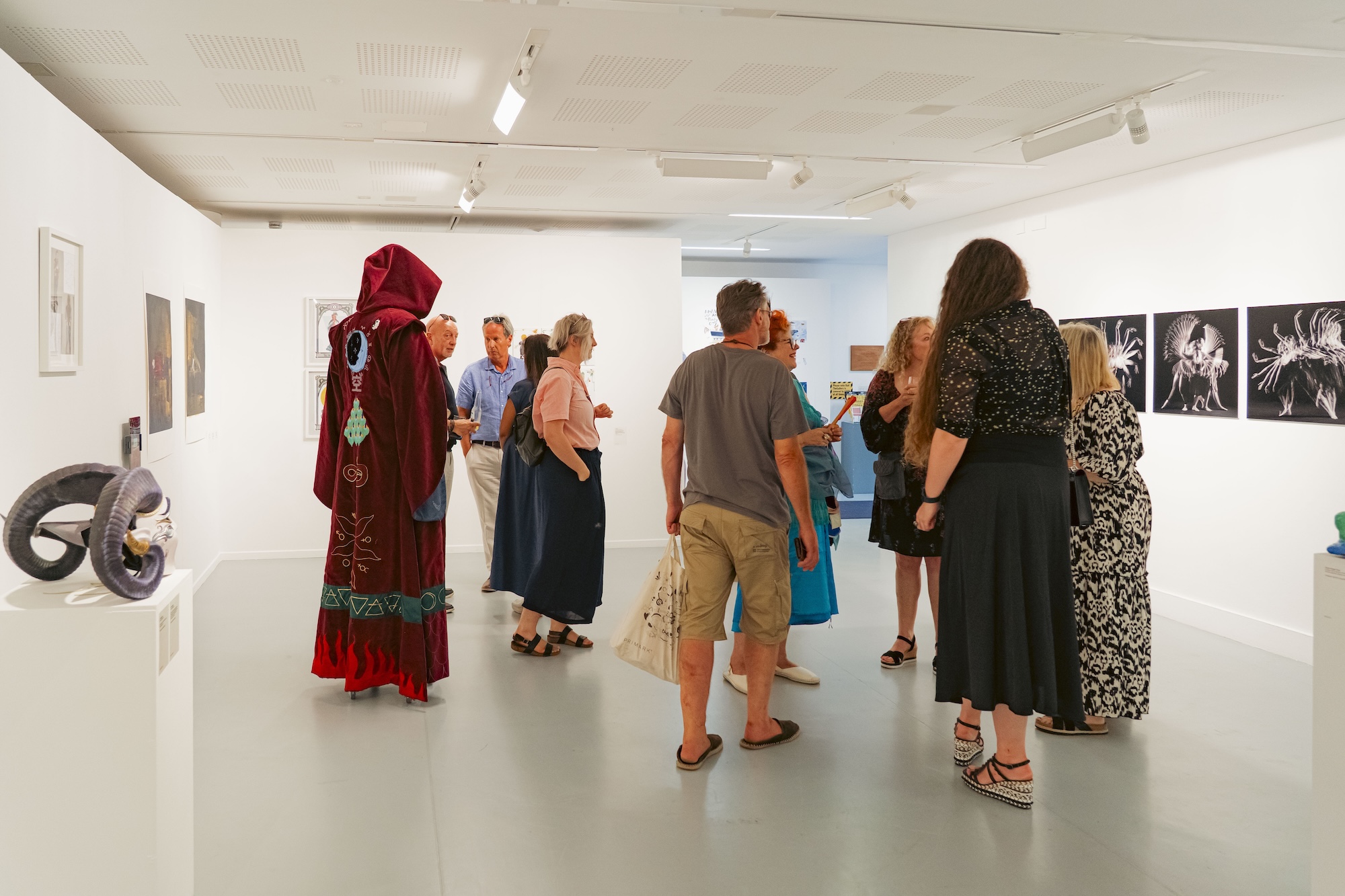 People stand and talk in a white-walled art gallery, with framed artworks on the walls and a red hooded costume-like figure among the group.