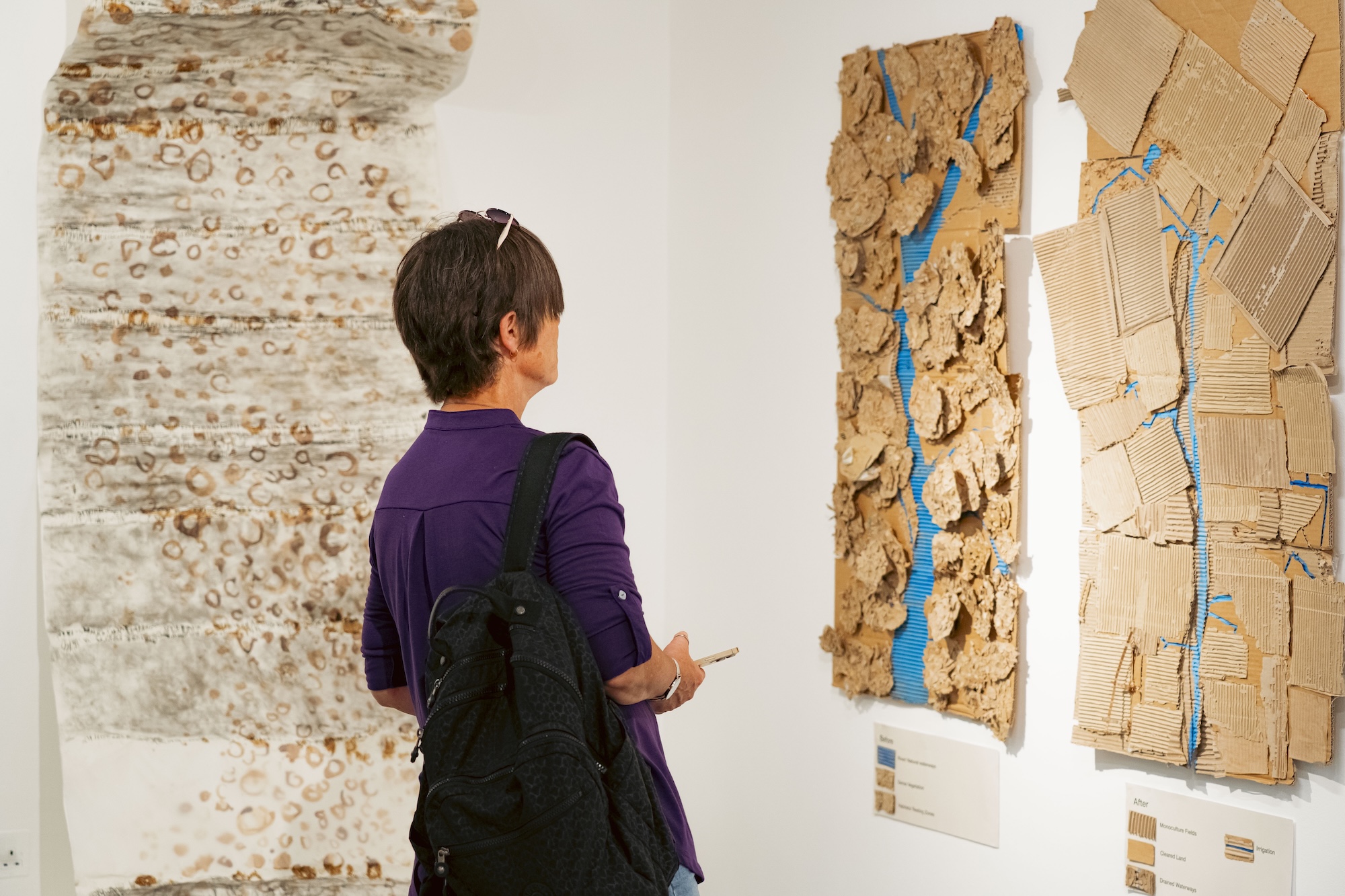 A woman with a backpack views three large abstract wall-mounted artworks in a gallery, including textured pieces with torn cardboard and blue accents.