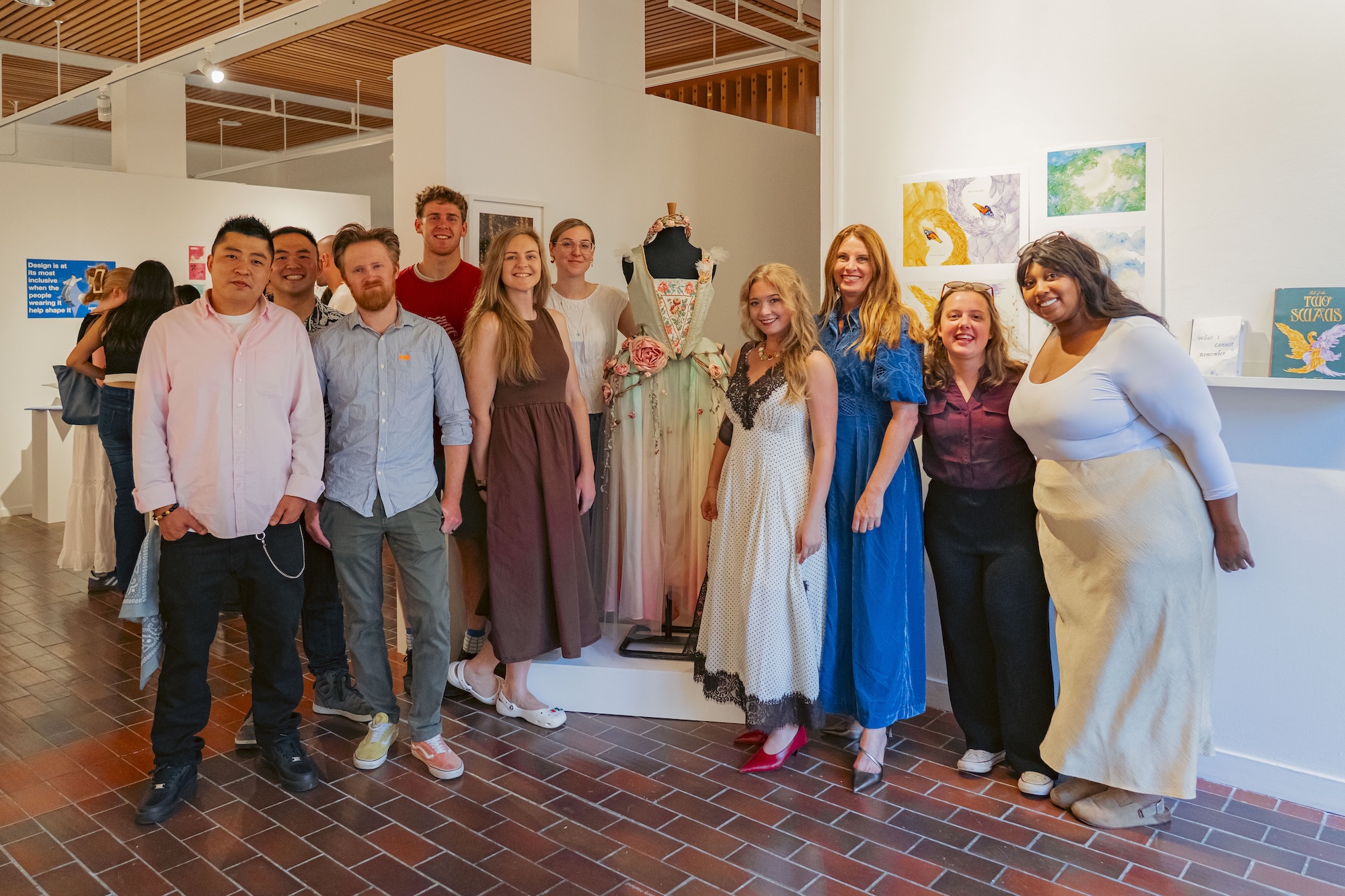 A group of people stands in an art gallery beside a floral dress on a mannequin, with framed artwork on white walls and a brick floor.