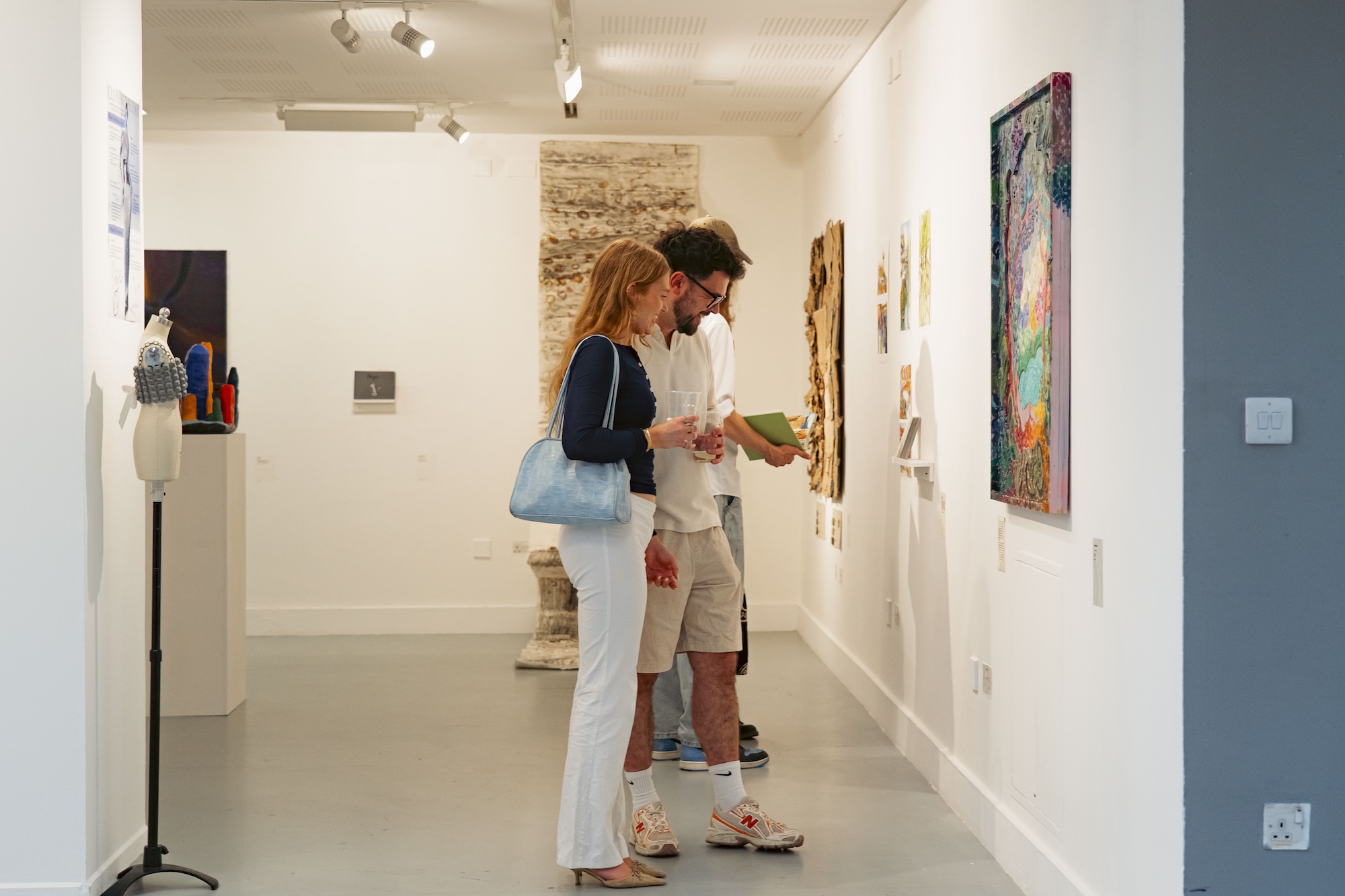 Two people viewing artwork in a white gallery hallway, with framed paintings on the walls and a display pedestal near the entrance.