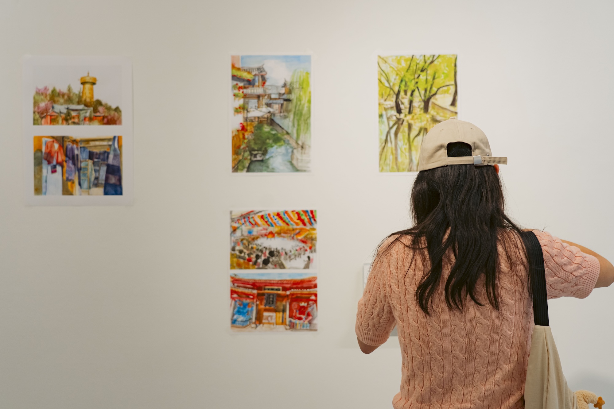 A woman in a beige cap and pink sweater studies small colourful paintings on a white gallery wall.
