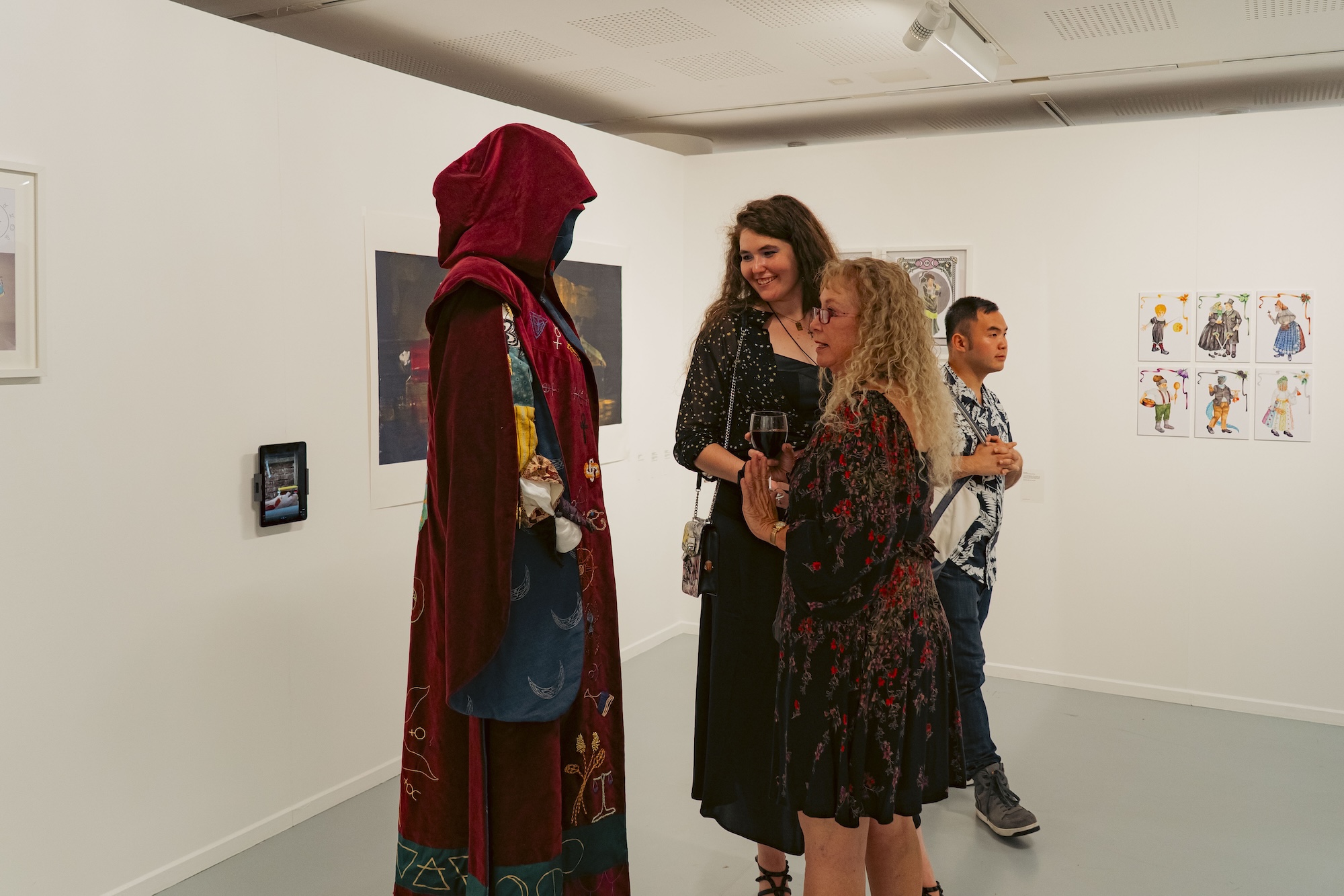 A hooded figure in a long red cloak stands facing three people in a white gallery, with framed artworks on the walls.