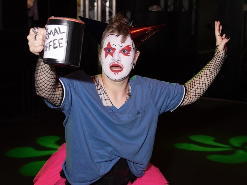 Person in white face paint with red details, blue shirt, fishnet sleeves, holding donation bucket whilst kneeling on pink mat.