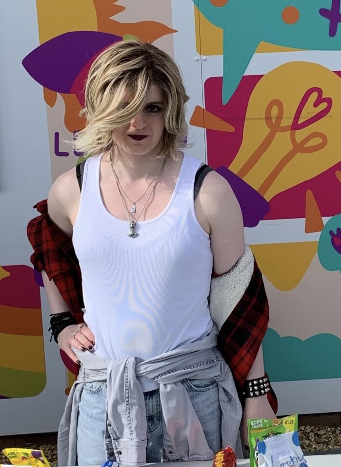 Woman with blonde hair in white vest and denim jacket against vibrant mural with purple, yellow, pink and teal abstract shapes.