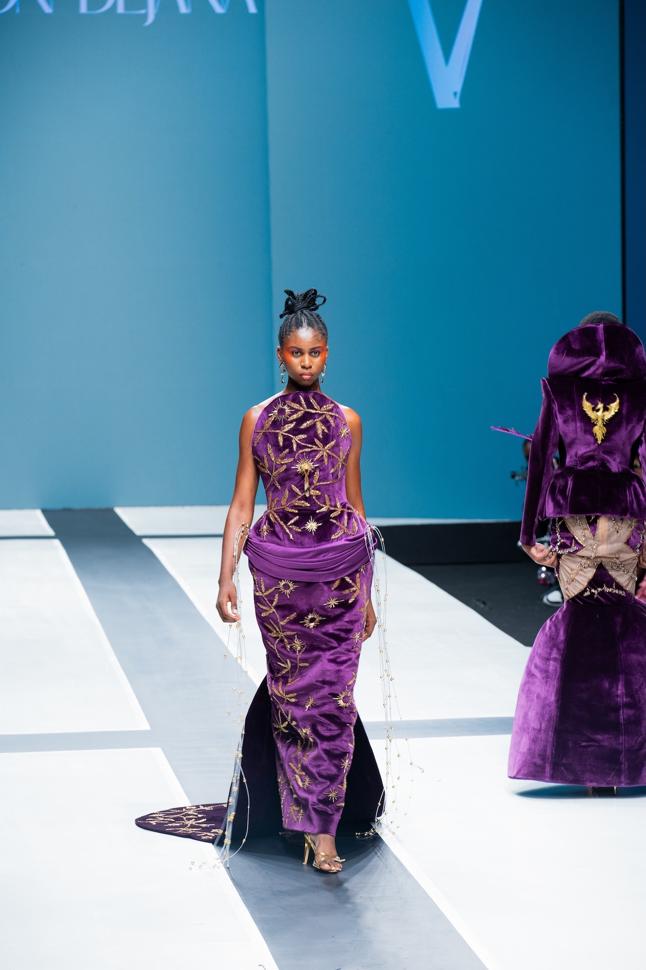 Model walking on white runway wearing purple dress with gold geometric cutouts, blue backdrop with partial white text visible.