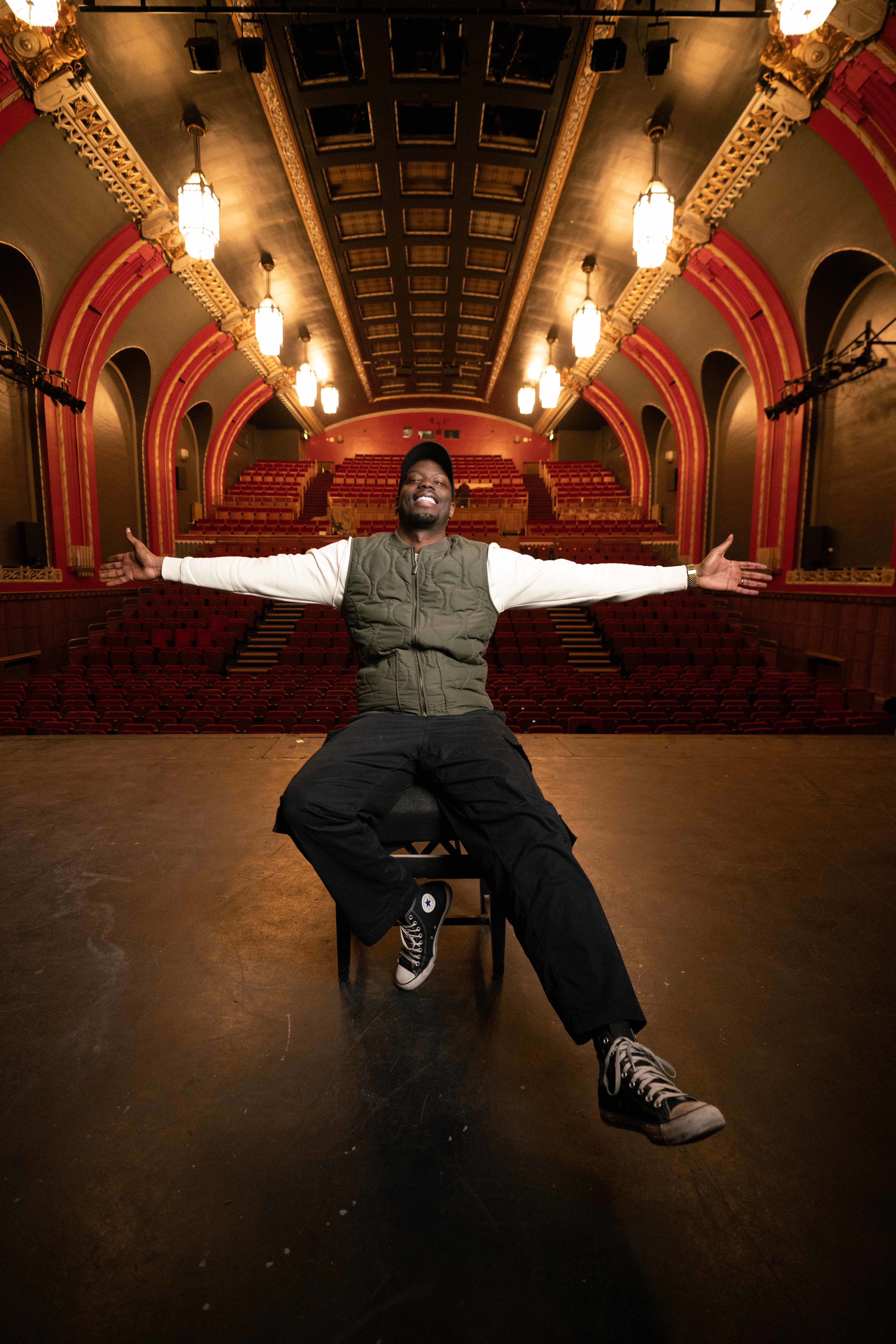 Ryan Calais Cameron sat in Lewisham Broadway Theatre on-stage with his arms extended out, smiling. Red archways an auditorium with chandeliers in the background.
