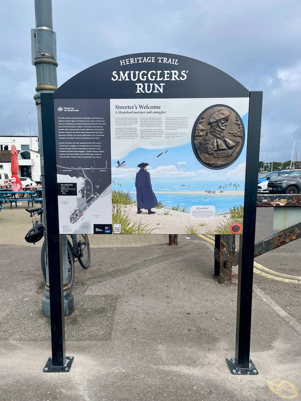 Heritage trail information board titled "Smugglers' Run" with text panels, bronze medallion, and illustration of cloaked figure by coastline.