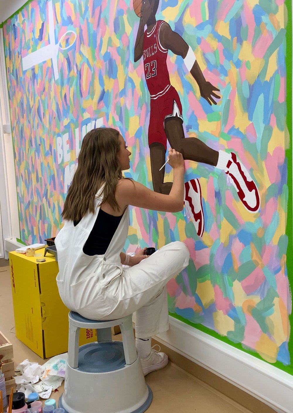 Tegan Price painting a person in basketball sportswear on a colourful background wall.