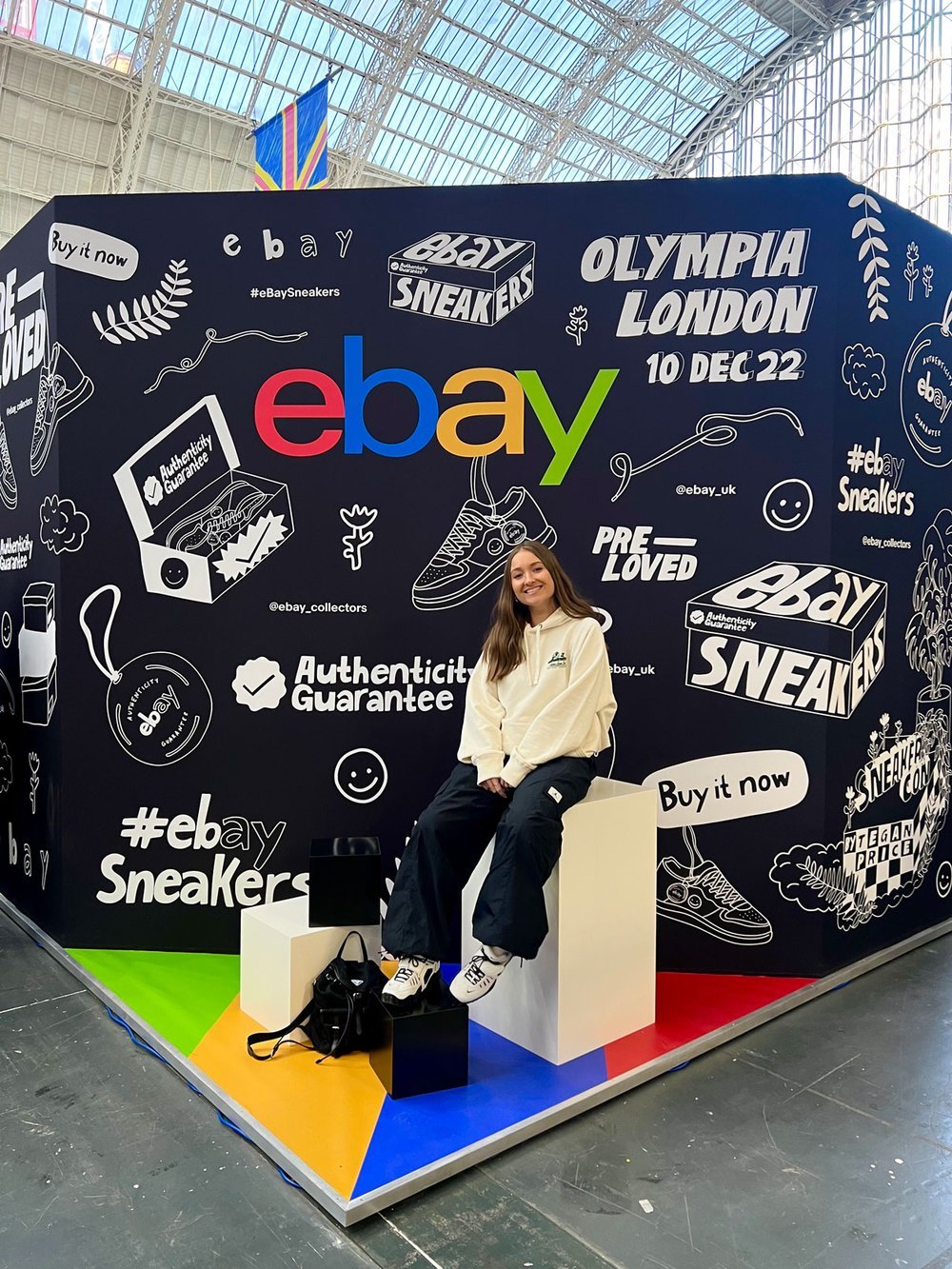 Tegan Price sat by a large ebay illustrated wall, with her illustration in white on a black background.