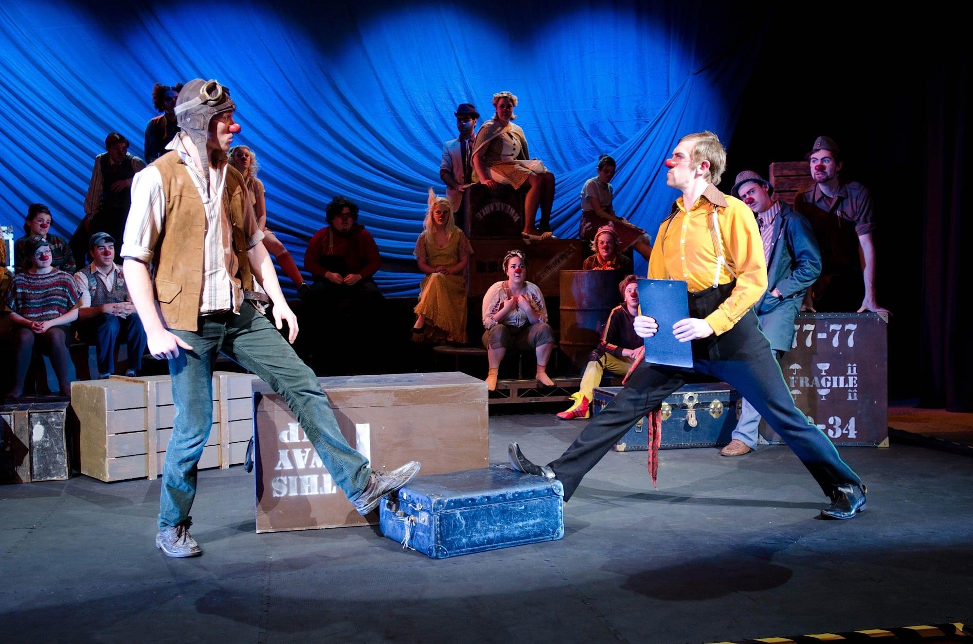 Two people with red noses stand opposite one another, each with a foot on a blue box on the floor in front of them.