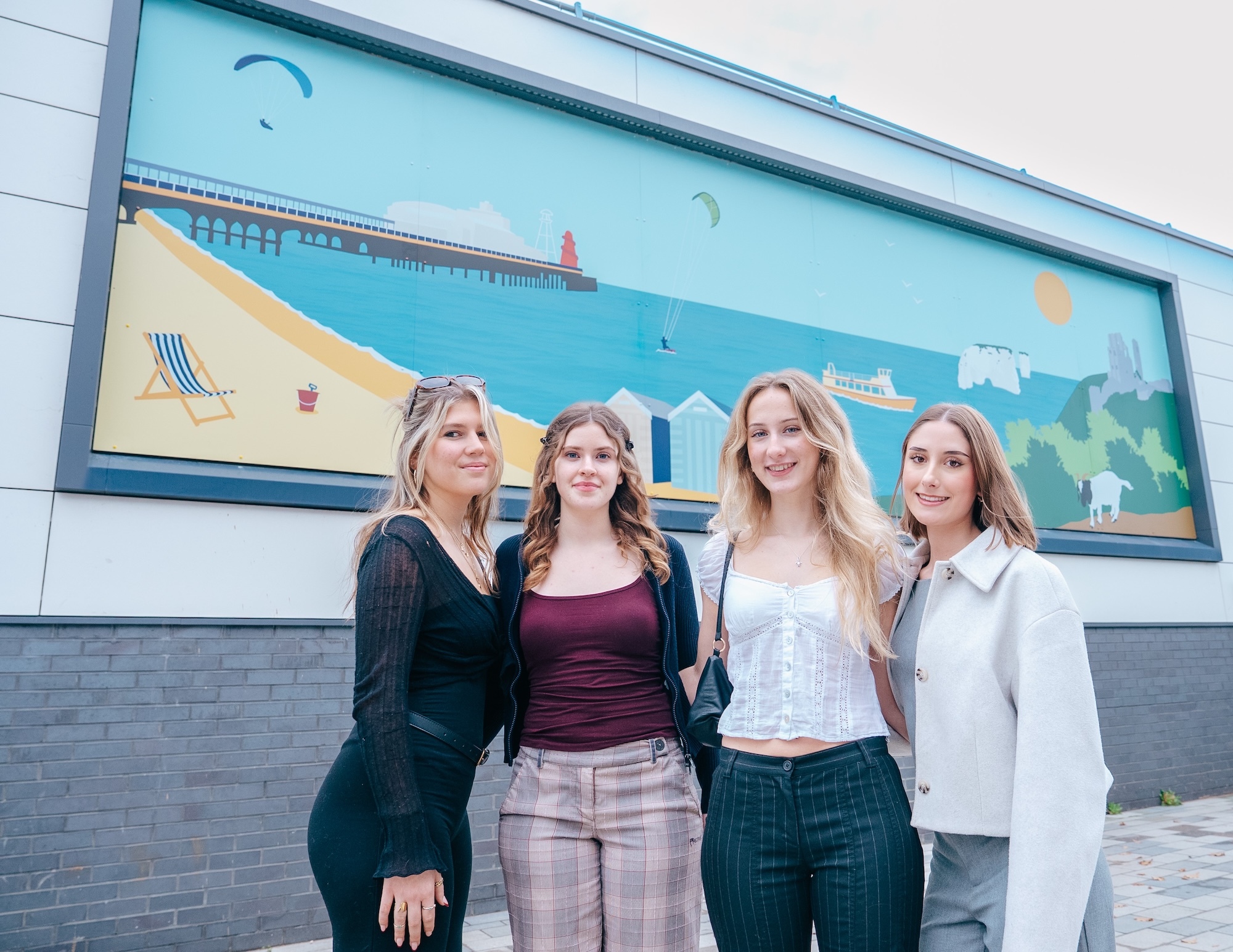 Four women standing together in front of a seaside-themed mural featuring blue sky, sandy beach with deckchair, and pier.