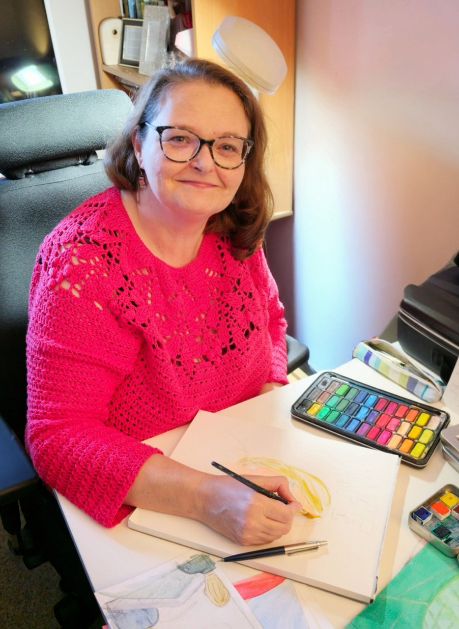A woman wearing glasses and a bright pink jumper sits at a desk, sketching with coloured pencils beside a tin of watercolours and an open drawing pad.