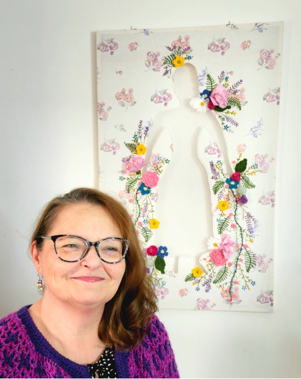 A woman wearing glasses and a purple crocheted cardigan stands beside a floral textile artwork mounted on a white wall.