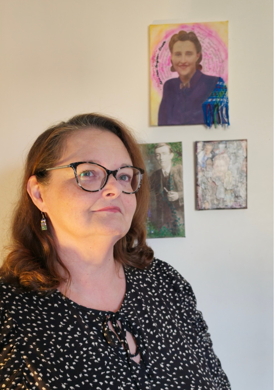 A woman wearing glasses and a black patterned blouse stands indoors beside three framed artworks on a white wall.