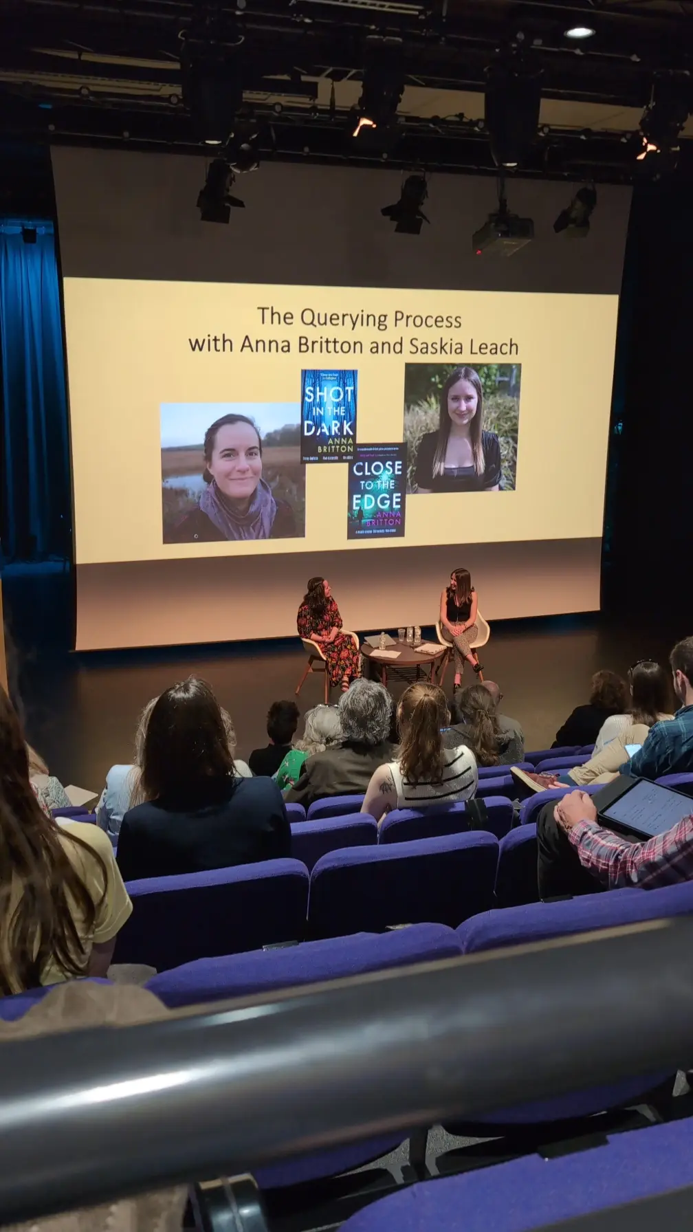 Two women speaking on stage in auditorium with blue seats. Large screen displays "The Querying Process with Anna Britton and Saskia Leach" and book covers.