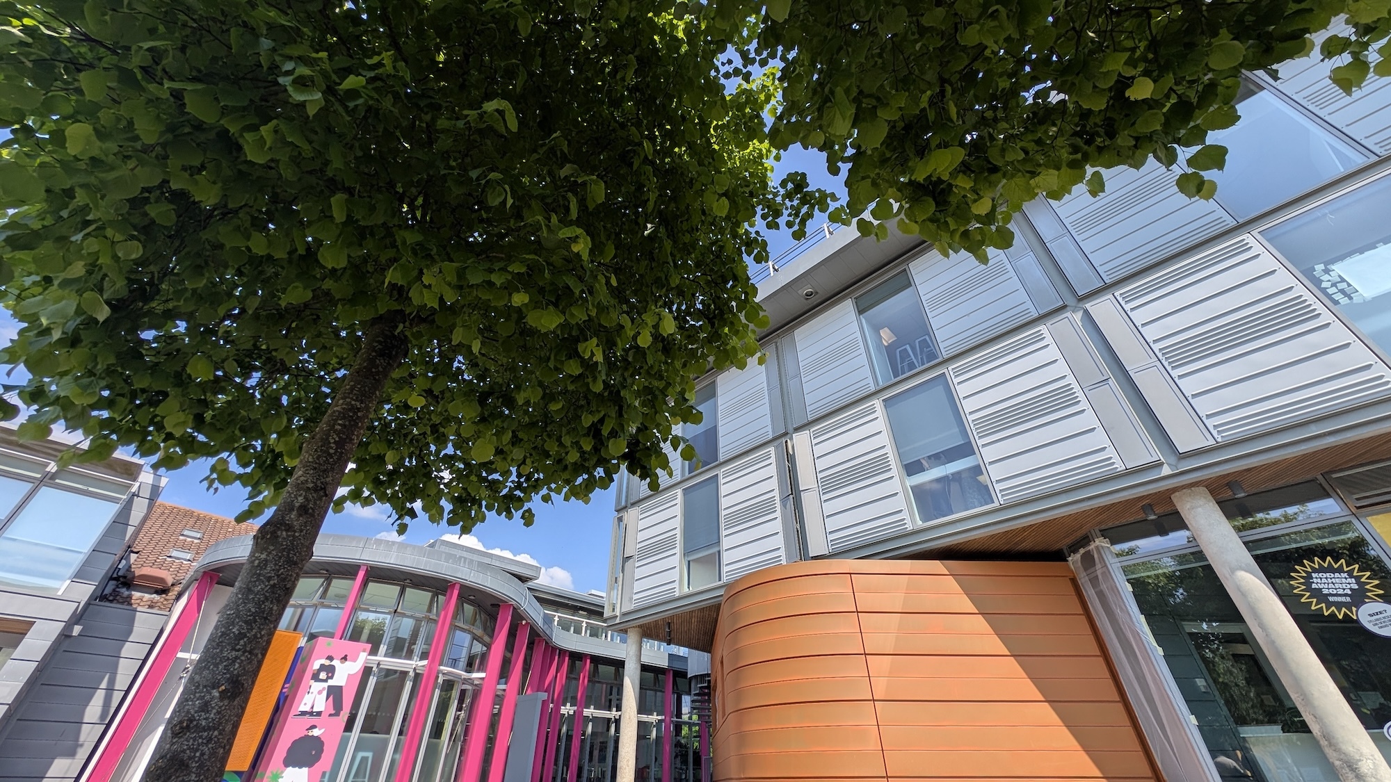 Part of Arts University Bournemouth – modern building with grey metal cladding and glass windows, orange-clad entrance section, green tree in foreground, blue sky above.