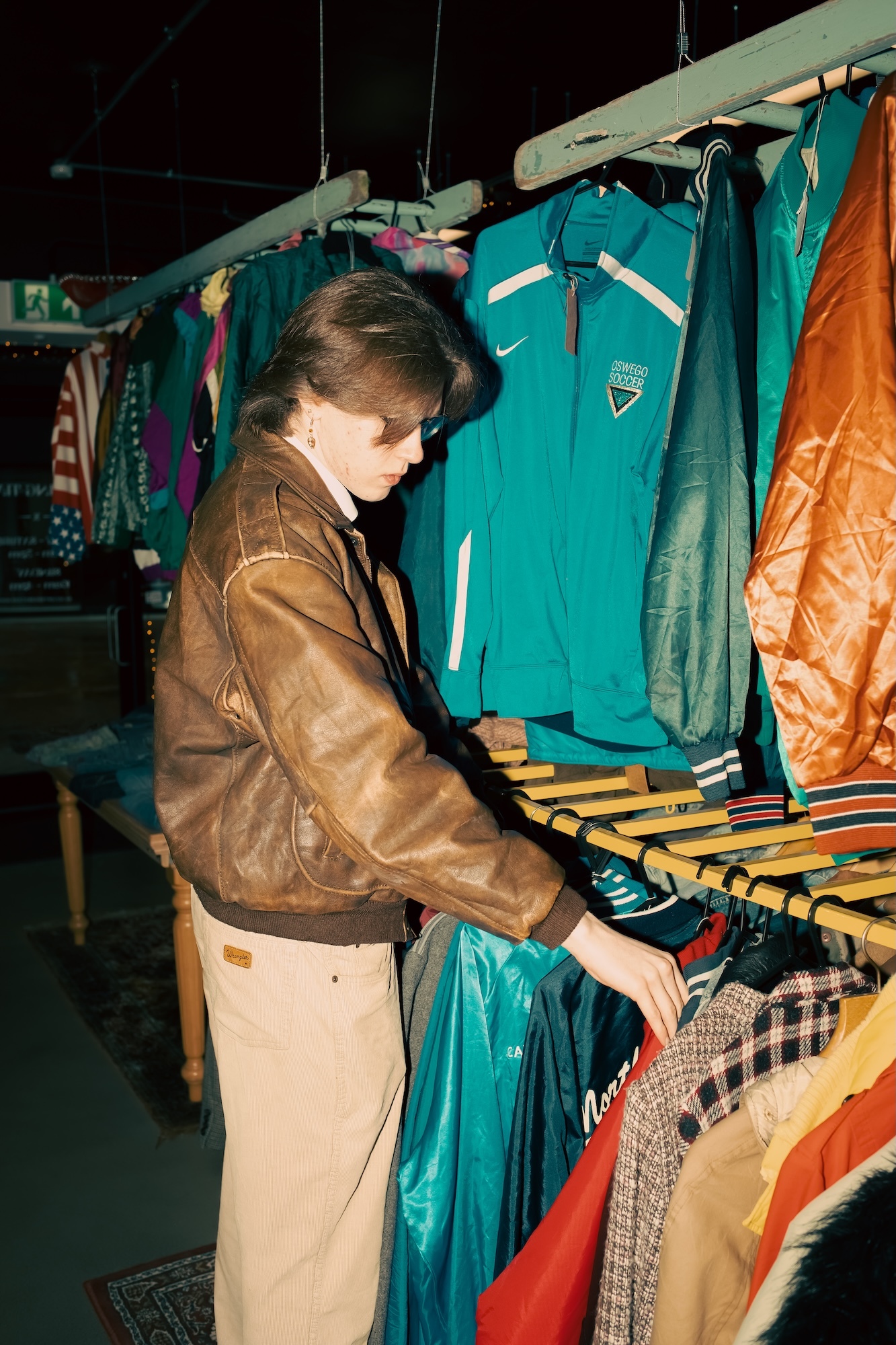 Person in brown leather jacket browsing clothes at market stall with teal tracksuits, orange jackets and colourful garments on hangers.