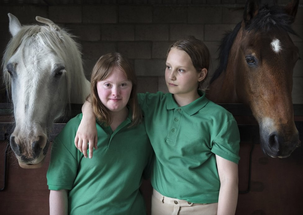 Two girls standing with their arms around each other in a stable. Two horses are stood either side.